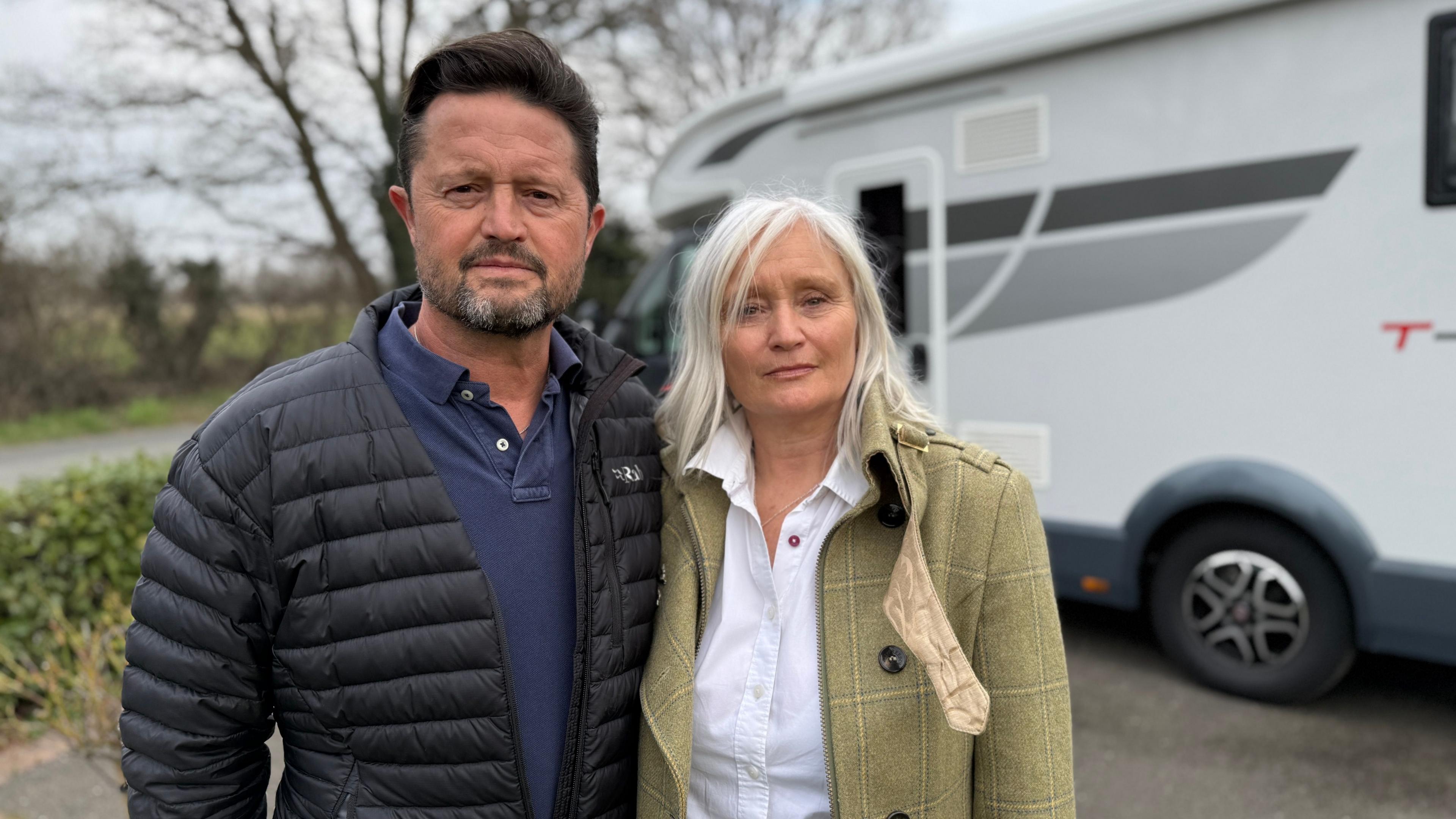 Adrian and Joanne Fenton standing acing the camera in front of their motorhome - he is wearing a black puffer jacket and she is wearing a pale green country wool coat