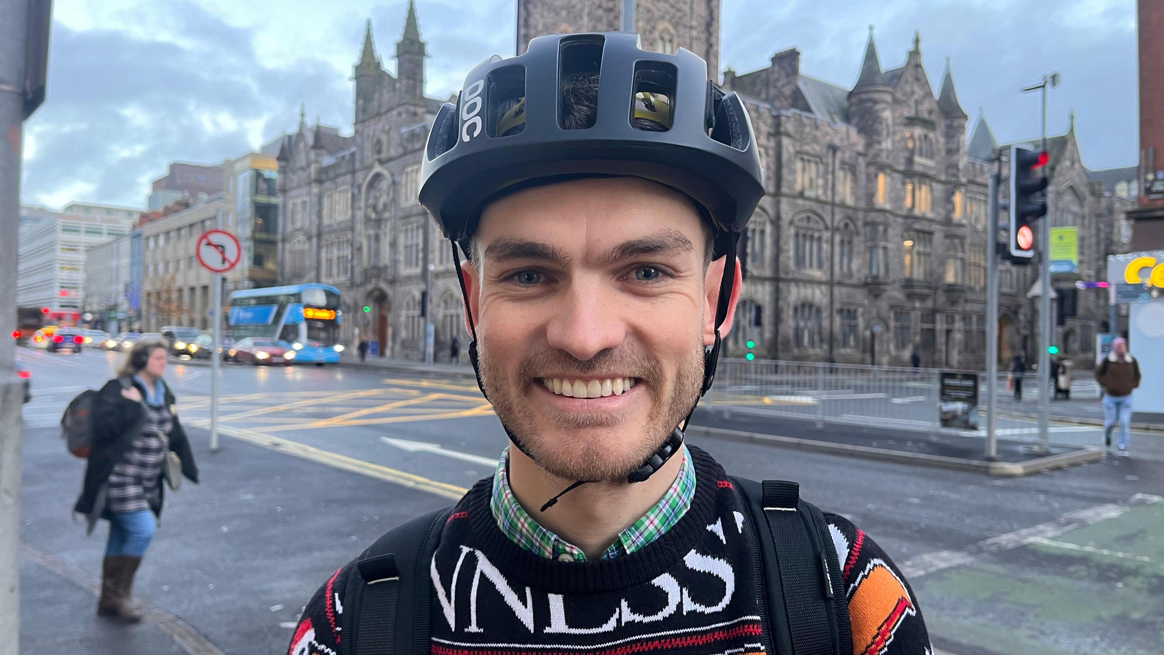 A man wearing a black bicycle helmet, he is wearing a black, white, red and orange jumper, with a green check shirt under it. 