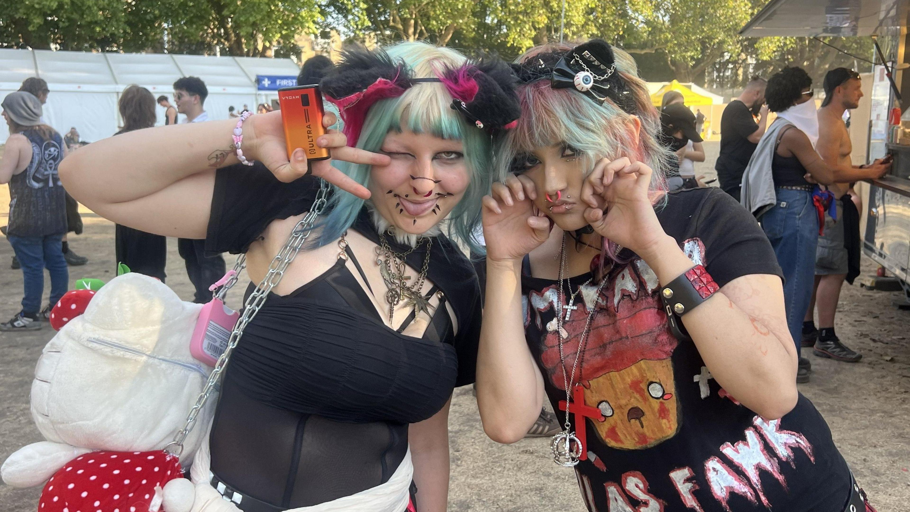 All Points East festival goers Bin and Zari grin astatine the camera wearing their festival gear. Bin wears a sheer achromatic apical and has greenish and blonde hairsbreadth with a achromatic furry headband. Zari wears a achromatic slogan t-shirt and has pinkish and greenish hair.