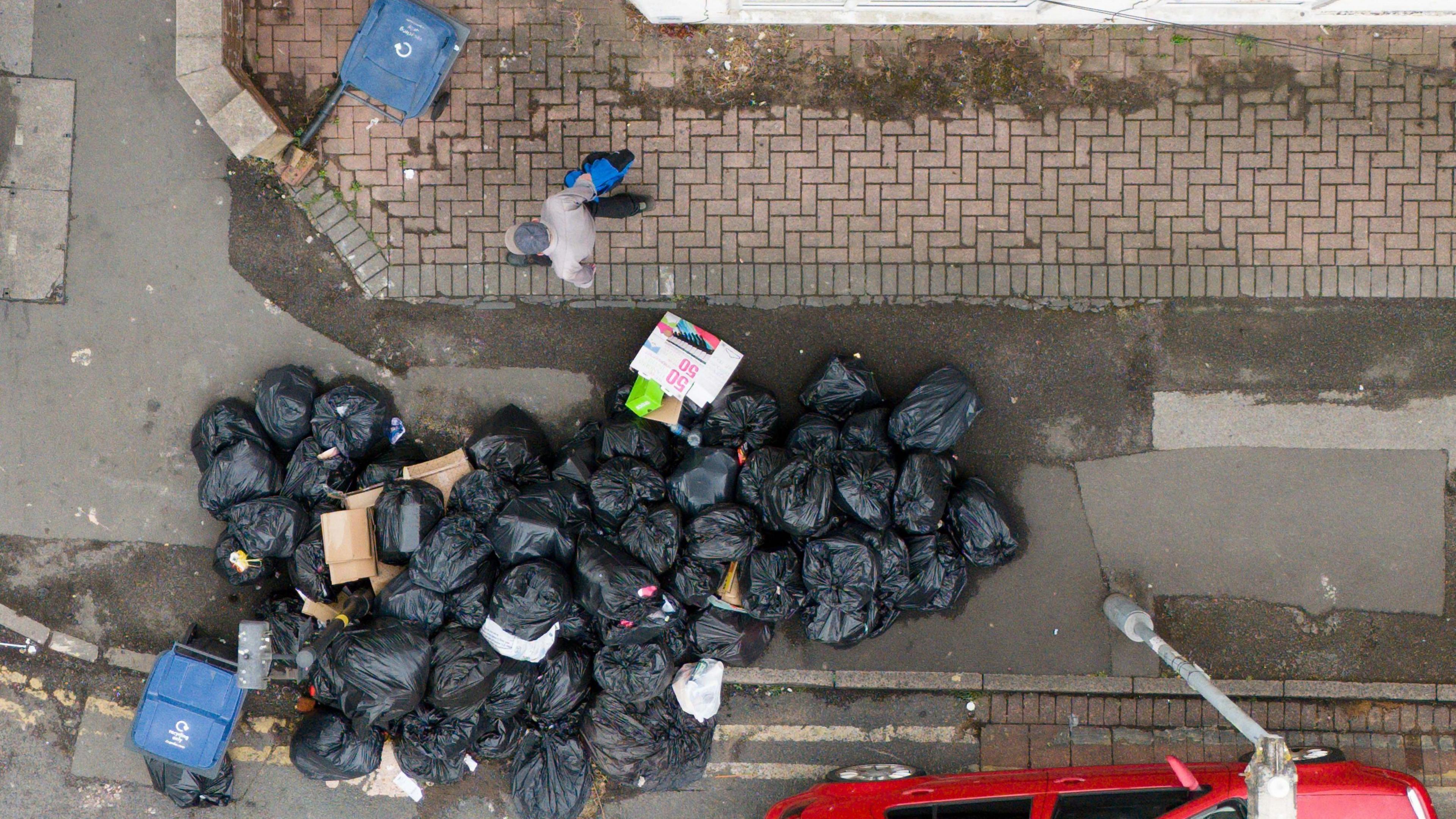 An aerial view of a large stash of bin bags spilling from a pavement into a road. A man walks past on the inside of the pavement. A blue bin can be seen next to the black bags, and dumped cardboard boxes.
