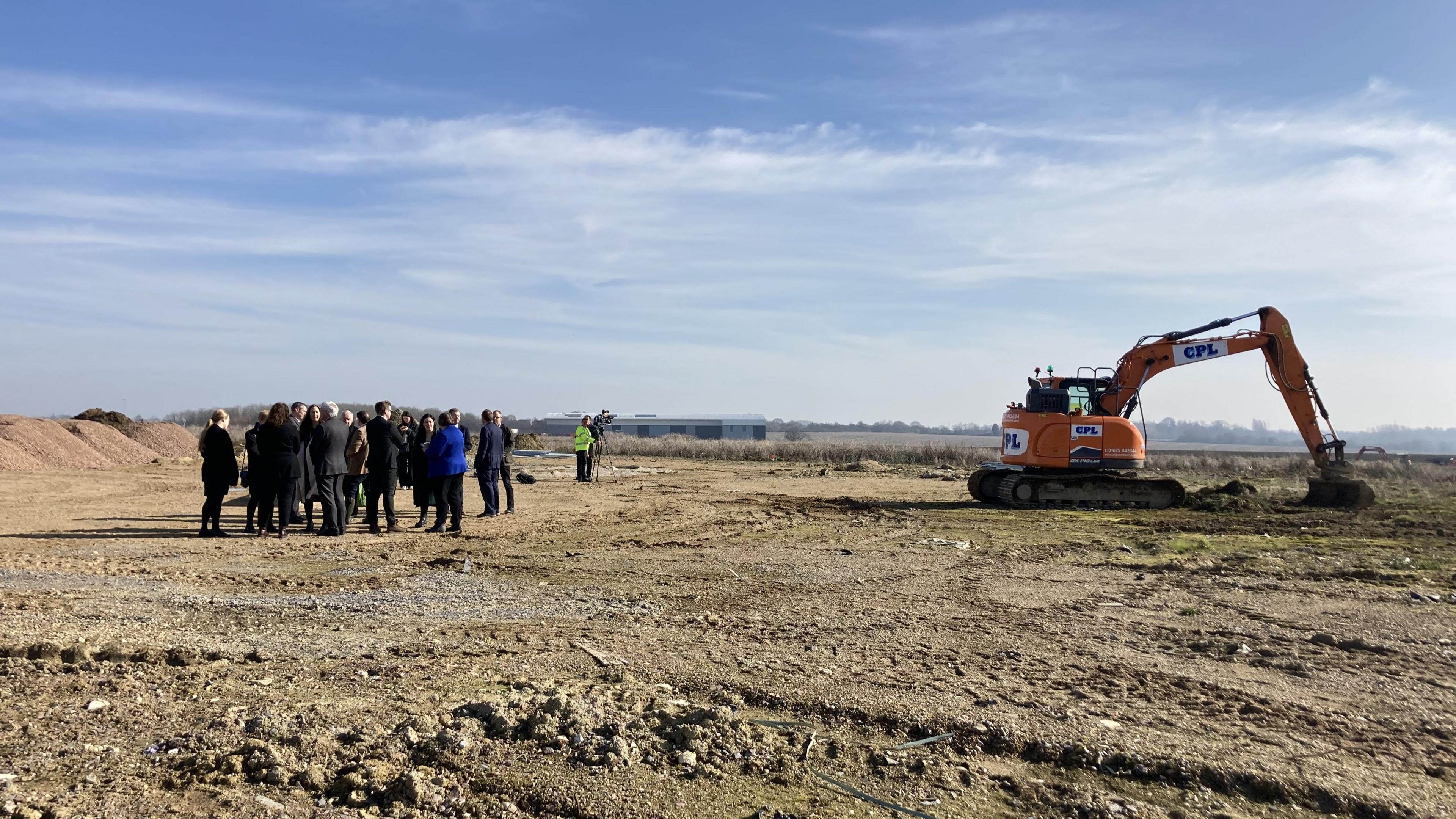 A group of people stand in a large field close to where a digger rests. 