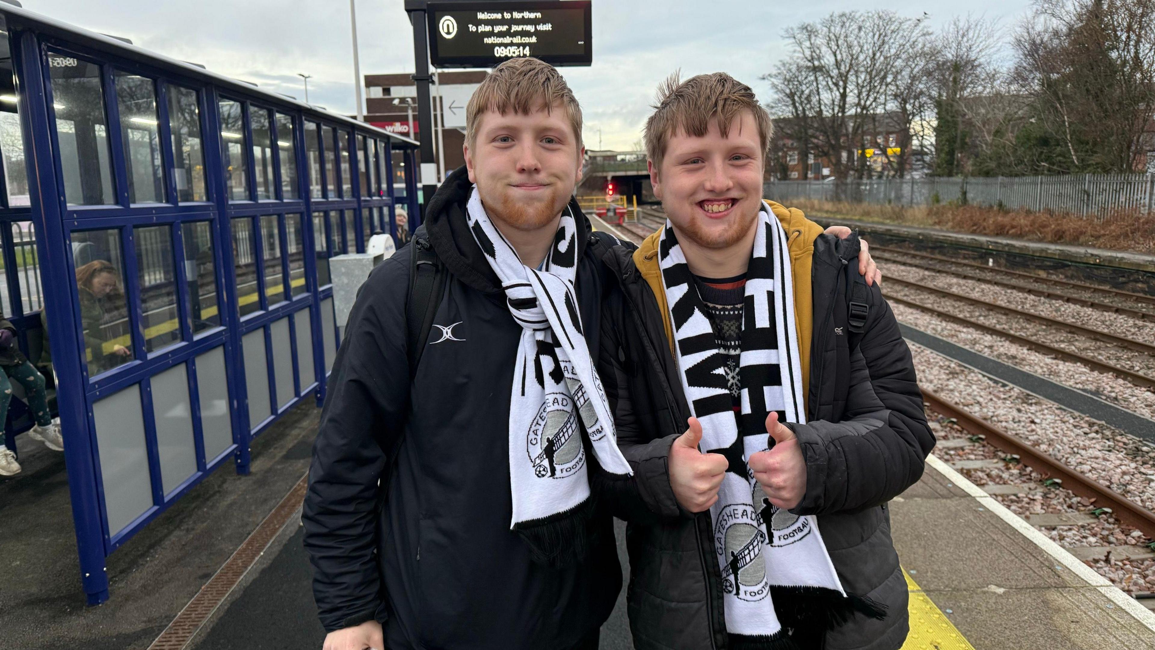 Twin brothers James and Thomas Lovejoy are wearing black and white Gateshead football club scarfs. They have light brown hair and smiling to the camera. They are standing on a station platform. 