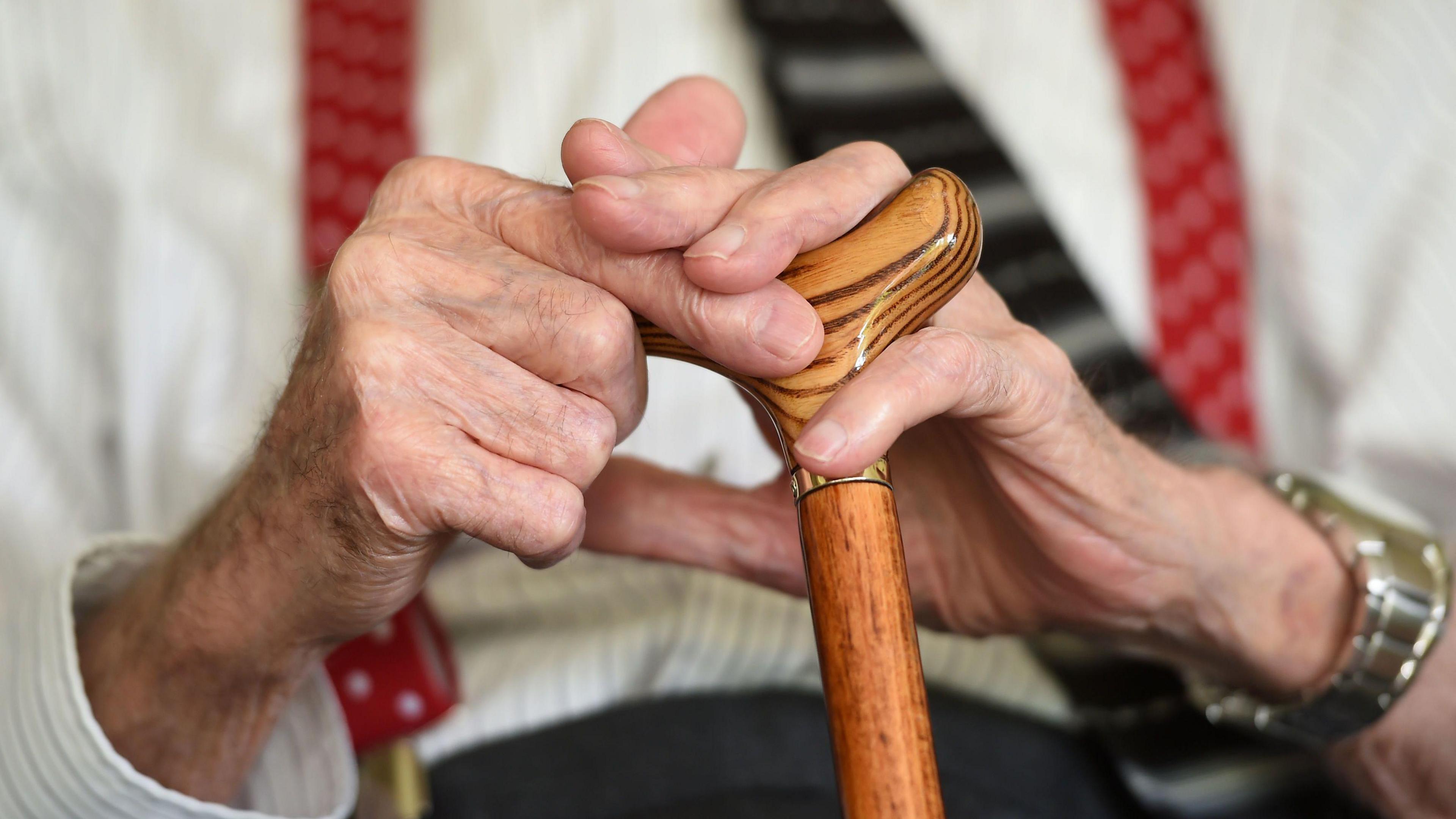 A close-up of hands holding on to the top of a light brown wooden walking stick. There is a watch on the person's right wrist and they are wearing a white shirt with red spotted braces. 