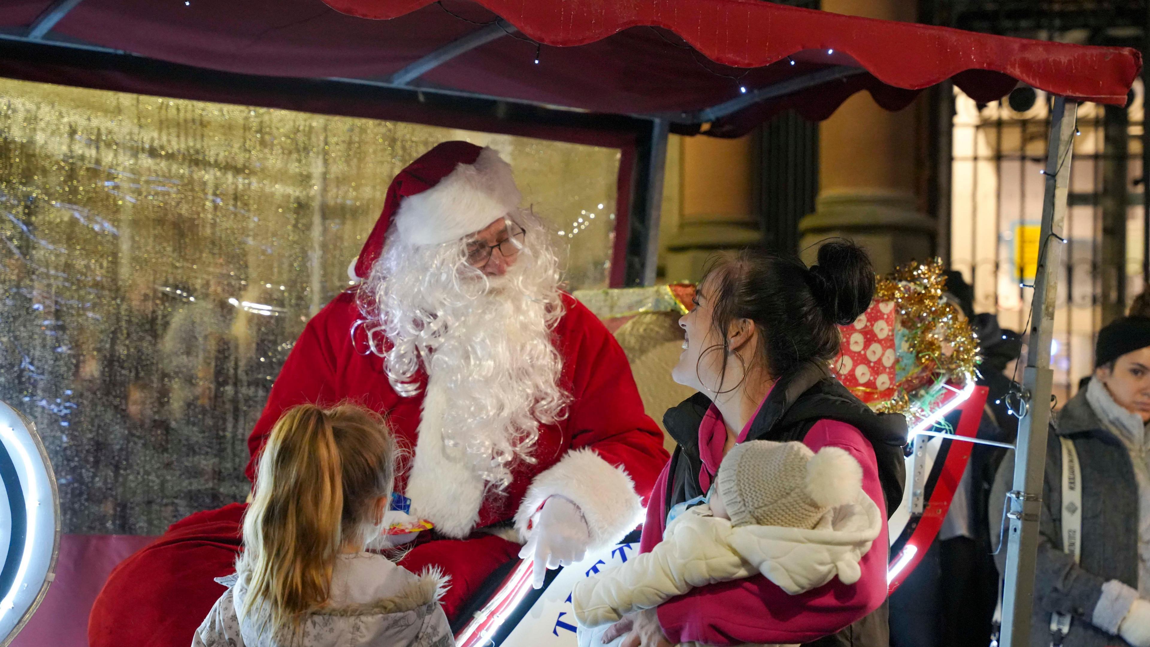 Father Christmas sitting in a sleigh with a present behind him talks to a woman holding a baby wearing a woolly hat with a bobble and a girl is standing to once side listening.