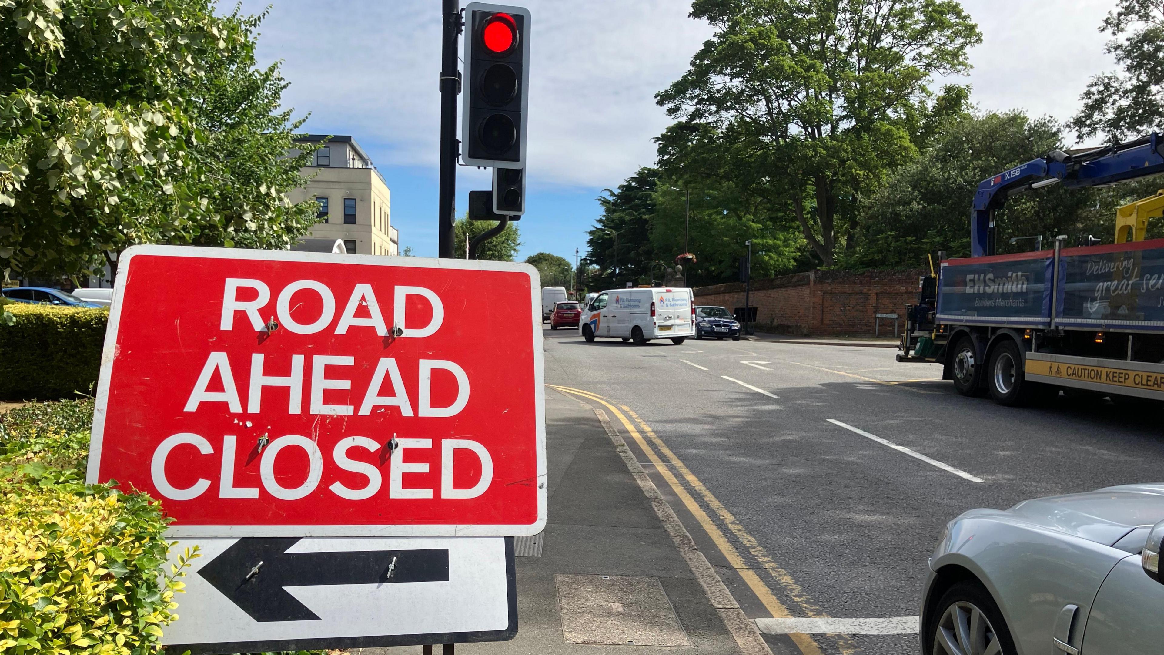 Red traffic sign indicating "Road ahead closed" at the side of a road in front of a red traffic light. A pavement hedge is on the left and a silver car is parked at the junction on the right. Vehicles can be seen crossing the junction.