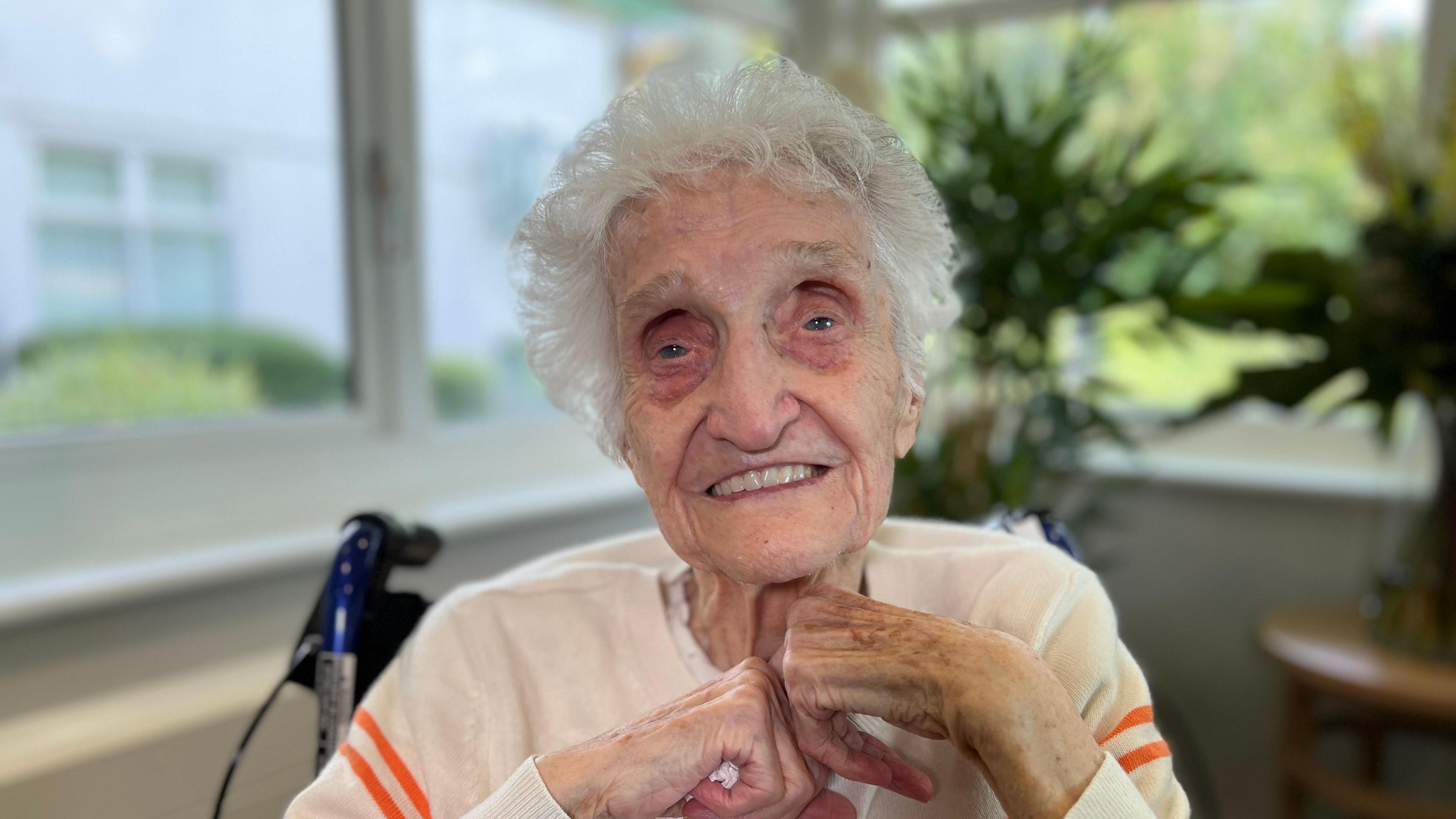 An elderly woman sits in a chair, wearing a white jumper with orange stripes and clasping their hands. Behind them are large windows showing greenery outside, along with some indoor plants