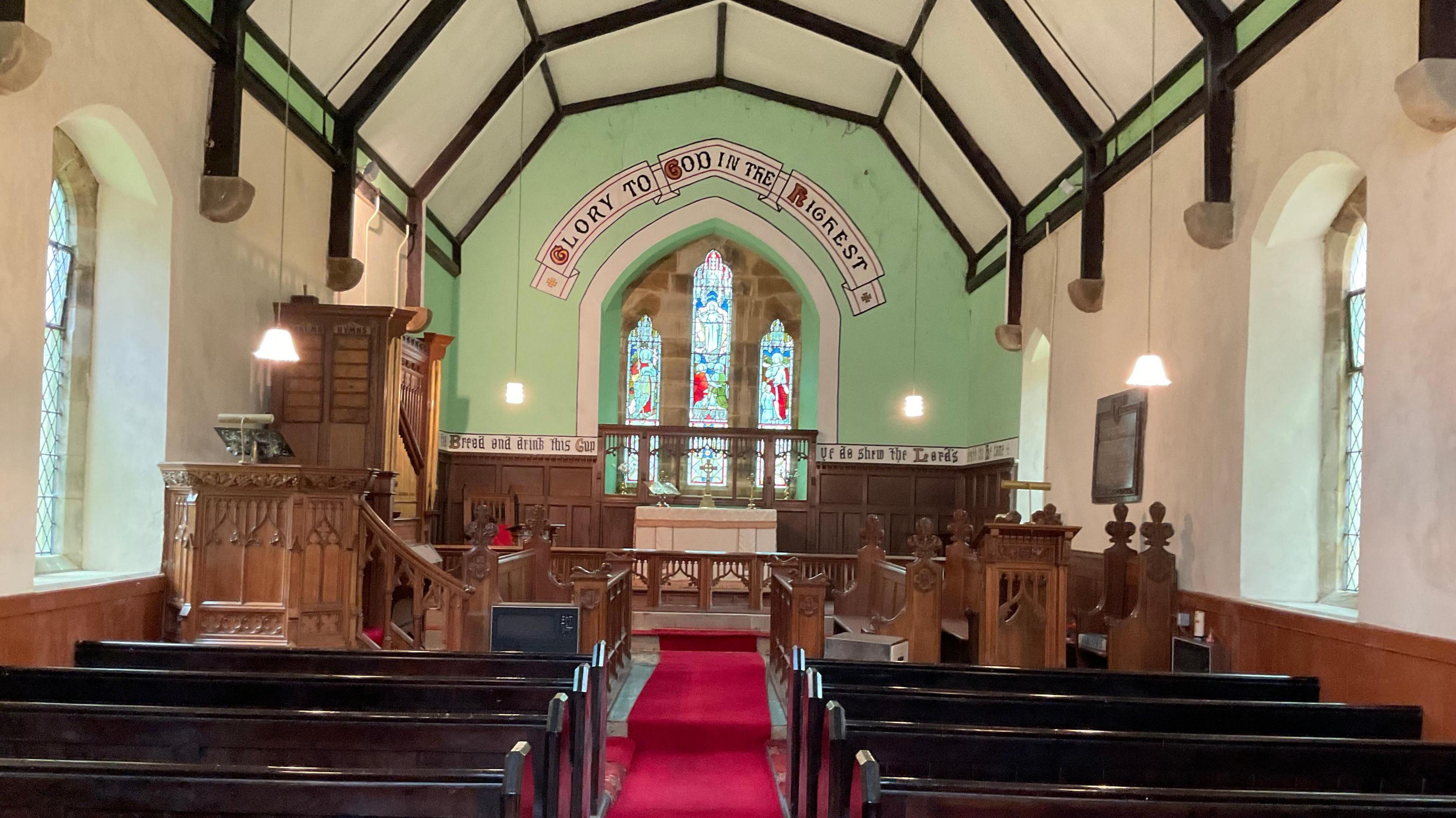 An interior view of All Saints Church. The pulpit stands to the left of the pews. A stained glass window is at the far end of the hall. Above it are the words: "Glory to god in the highest".