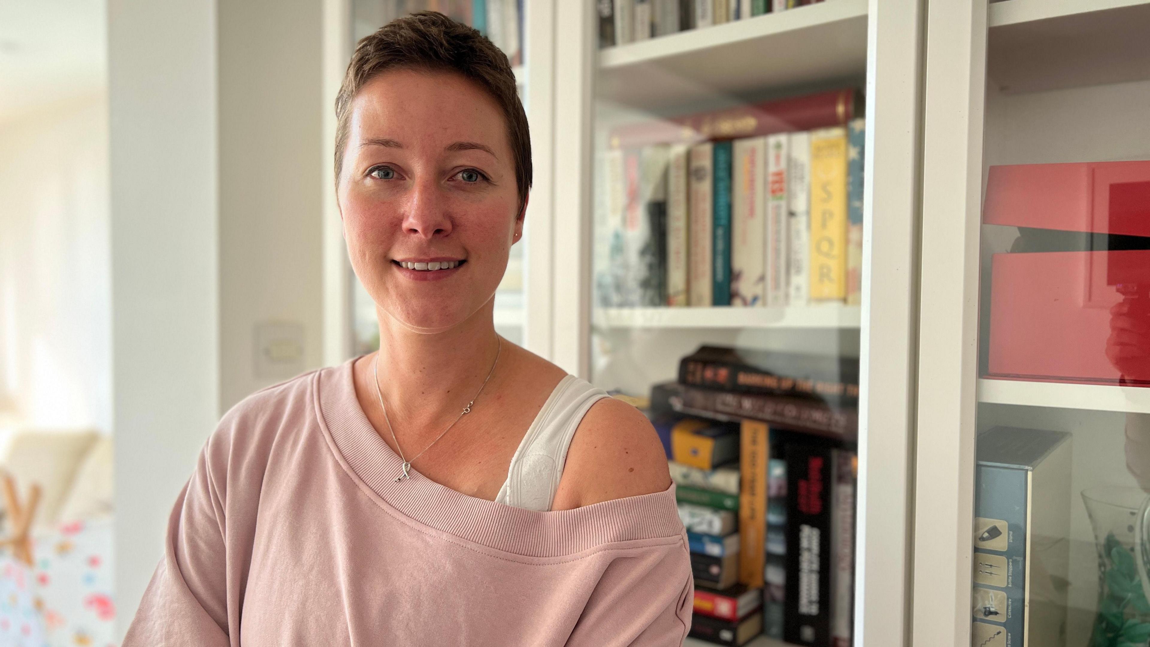 Margi looks at the camera and smiles. She is wearing a pink jumper with her right shoulder showing. She has short brown hair and blue eyes. She's standing in front of a book case.