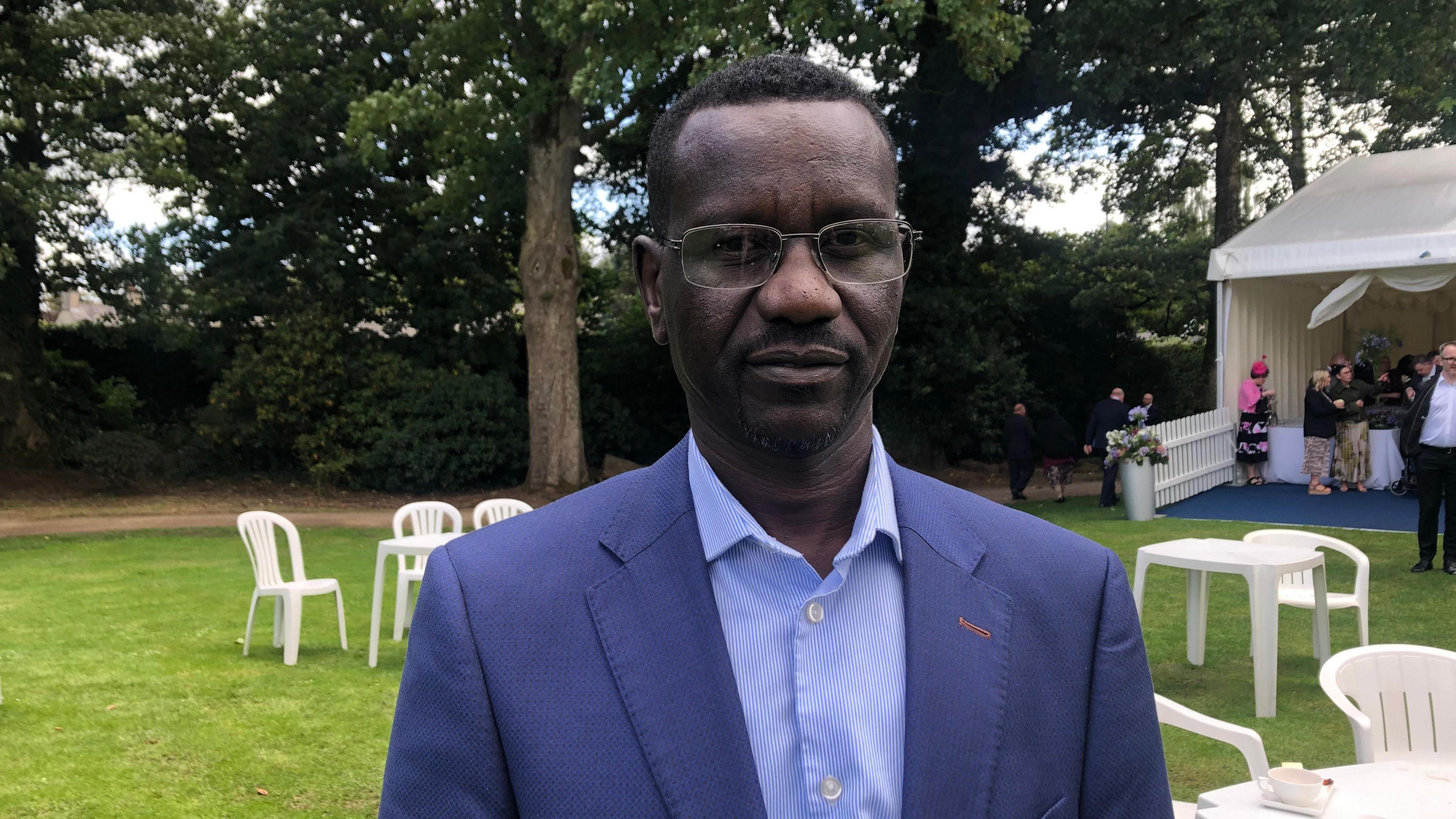 Man standing in a navy blue suit and navy shirt standing on grass. 