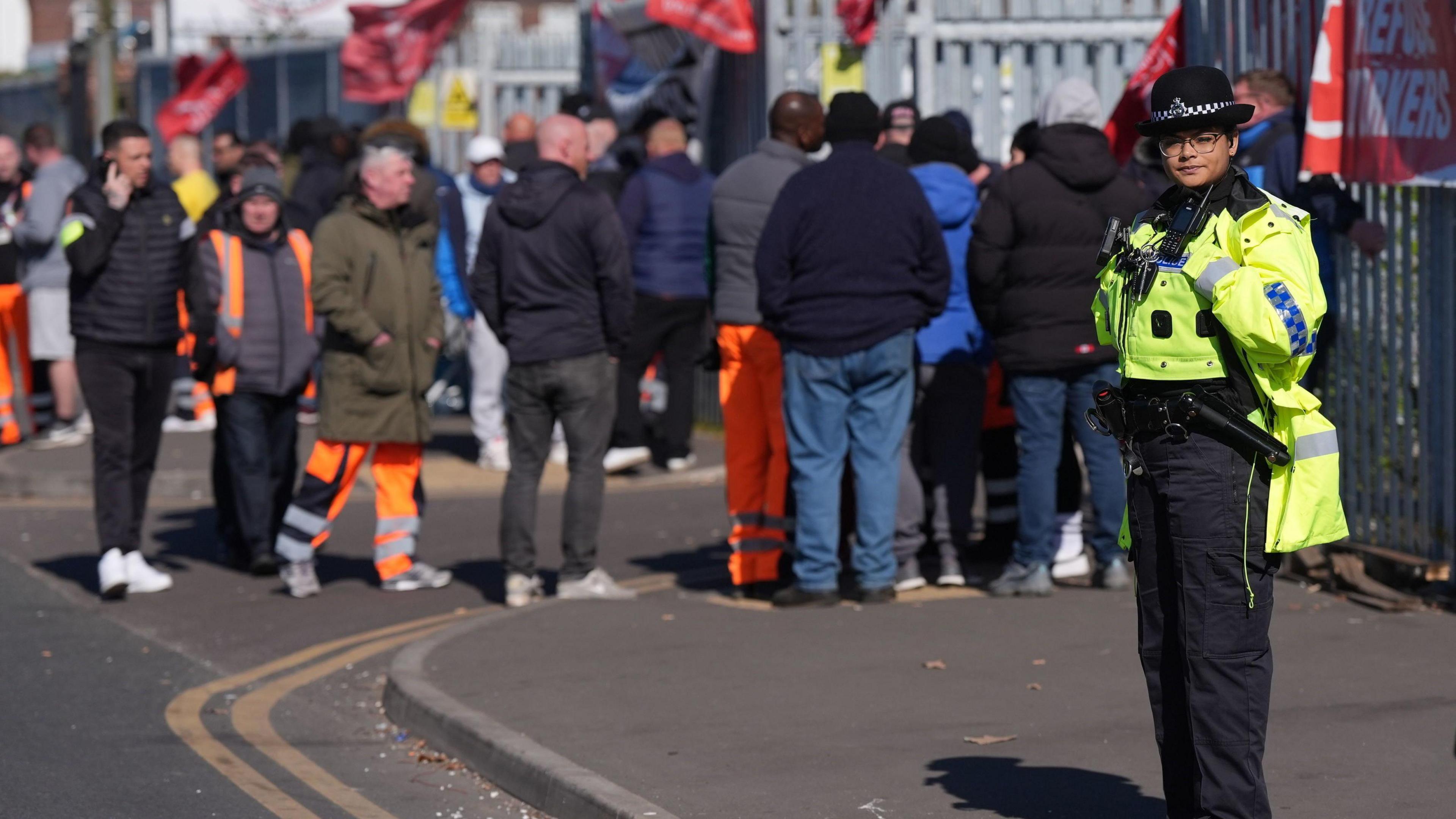 A police officer stands in the foreground on a pavement, looking in our direction. Behind her is a number of people on a picket line in front of the large metal gates of a waste facility.