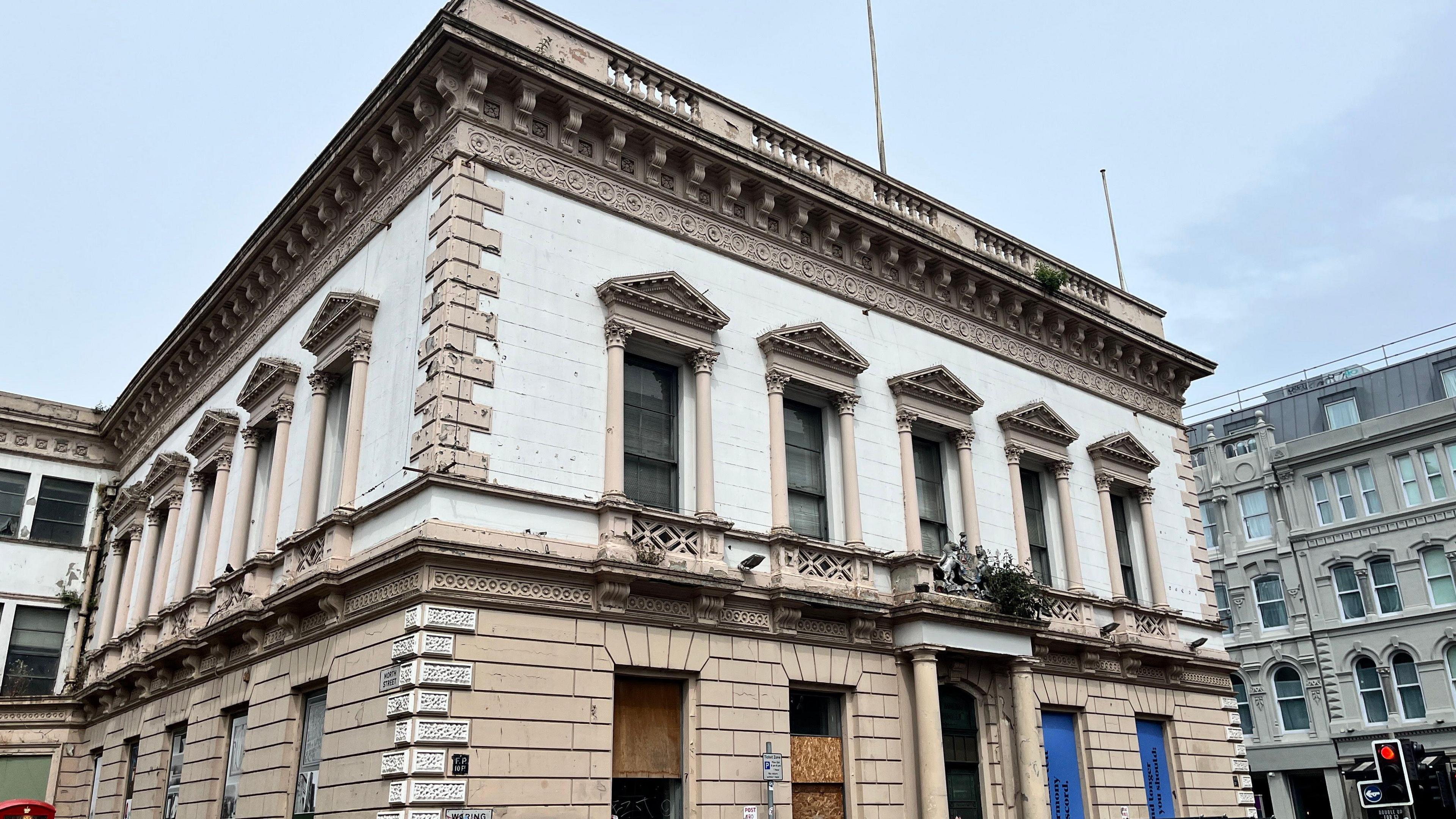 A large square building in Belfast. The building is white and light brown. The windows are boarded up, some are brown and some blue. In the background there are other large buildings, they are grey. In the corner is a traffic light on red.