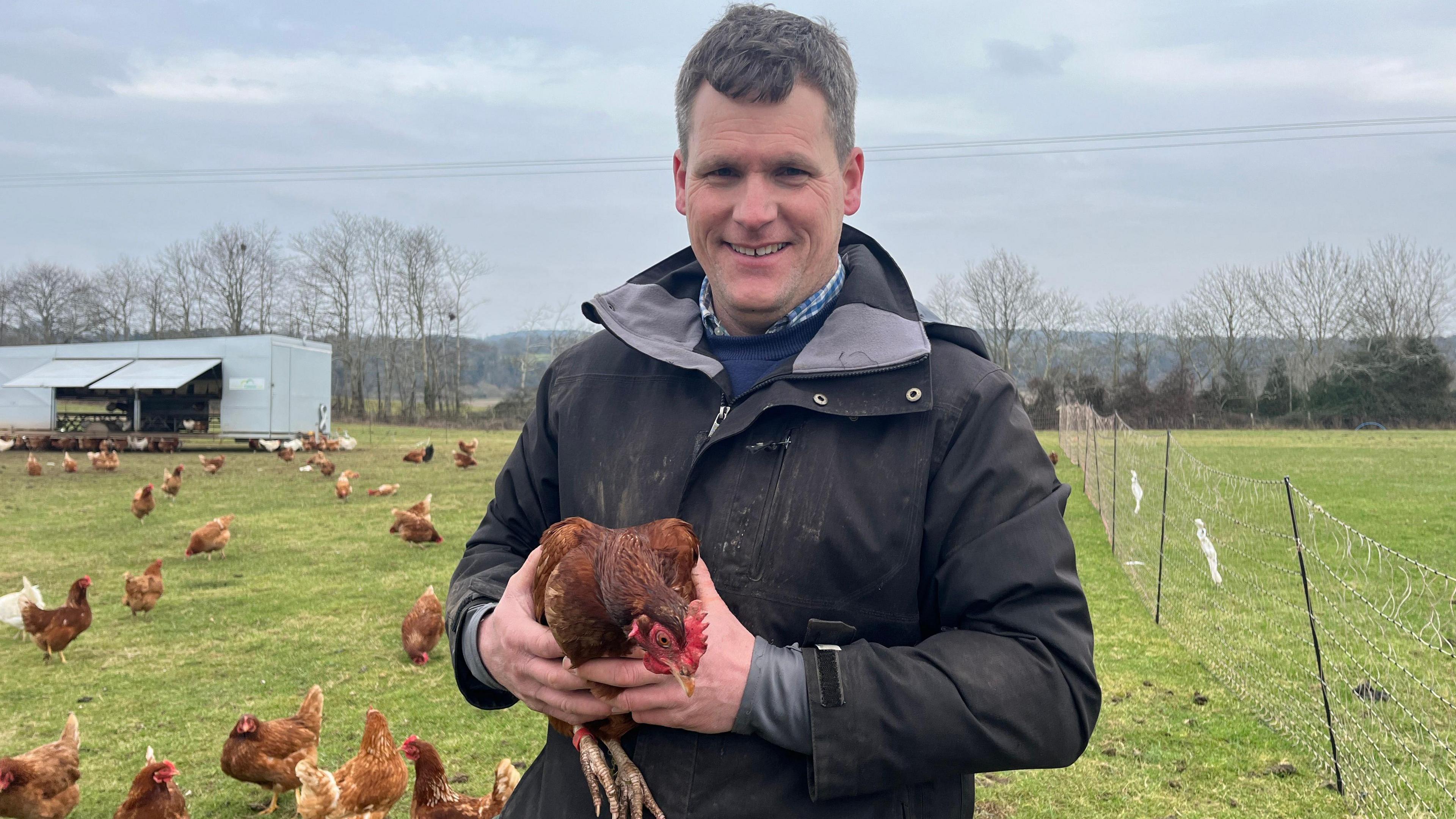 A man with a black jacket stands holding a hen. Other chickens can be seen in the background on the grass.