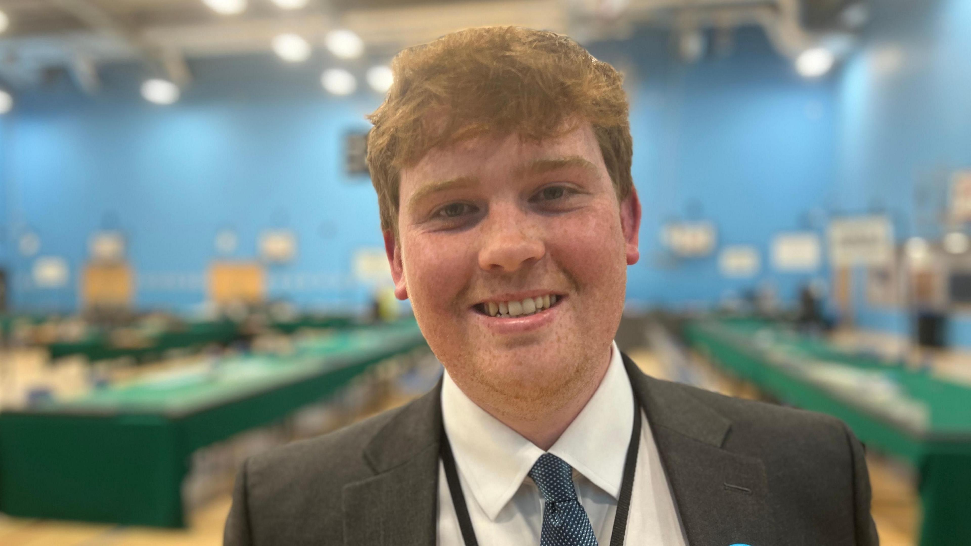 A man with medium-length light brown hair, smiling at the camera and wearing a dark jacket, a white shirt, a blue tie and a lanyard. He is at an election count and tables covered in green cloths are behind him.