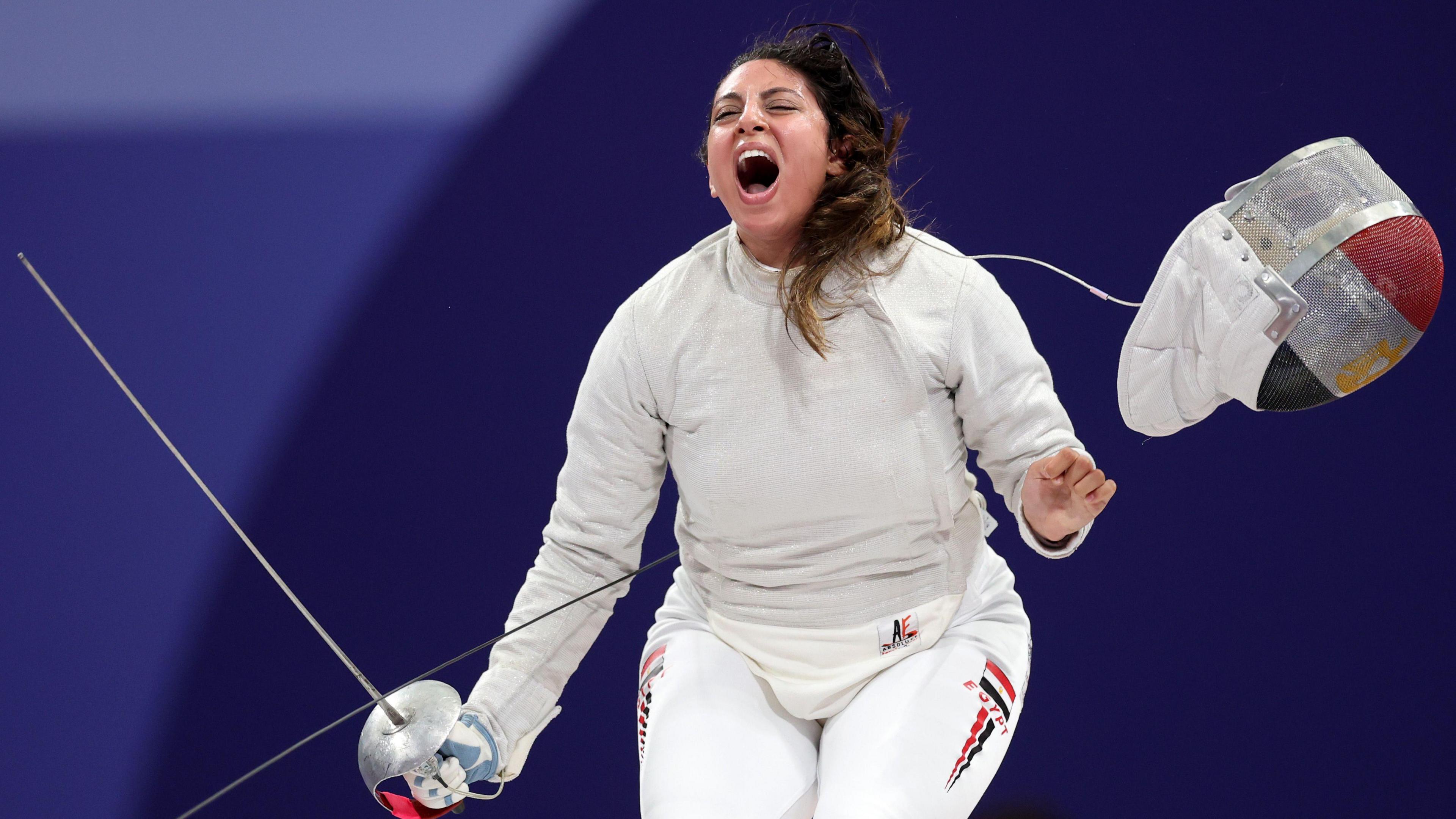 Nada Hafez, wearing a white fencing uniform and holding a sabre in her right hand, lets out a scream of celebration after throwing off her protective mask bearing a design of the Egyptian flag