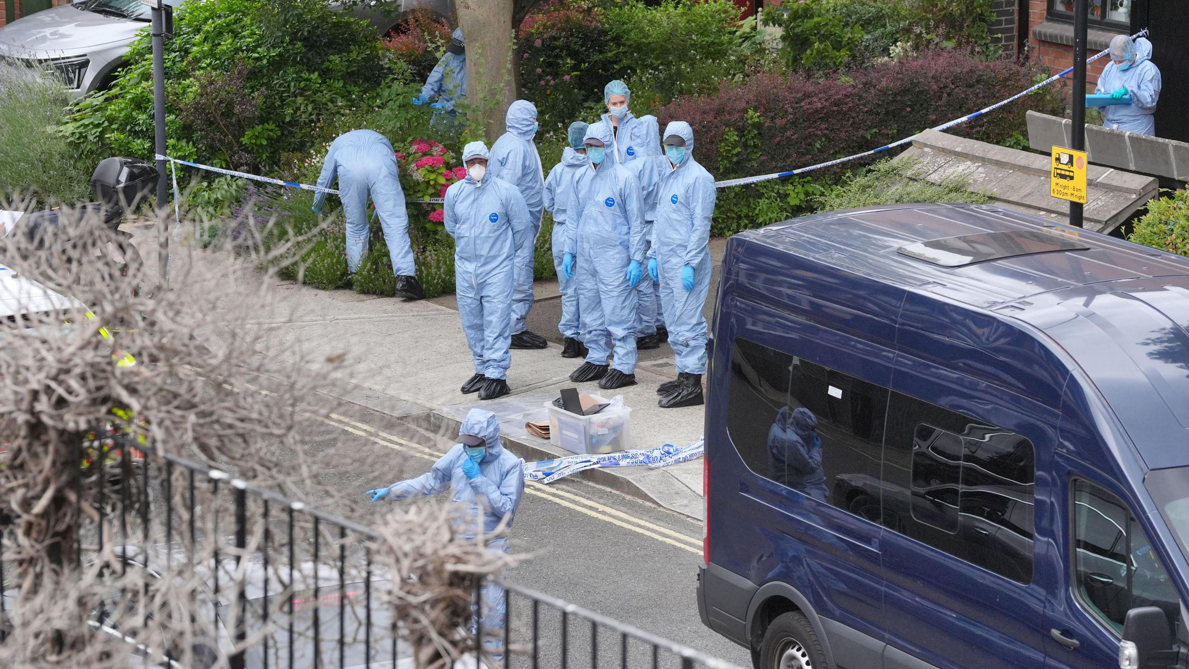 A group of forensic investigators dressed in light blue hazmat suits outside of the crime scene in Shepherd's Bush, London. There is a dark blue van in the image and blue and white forensic tape blocking access to the property.