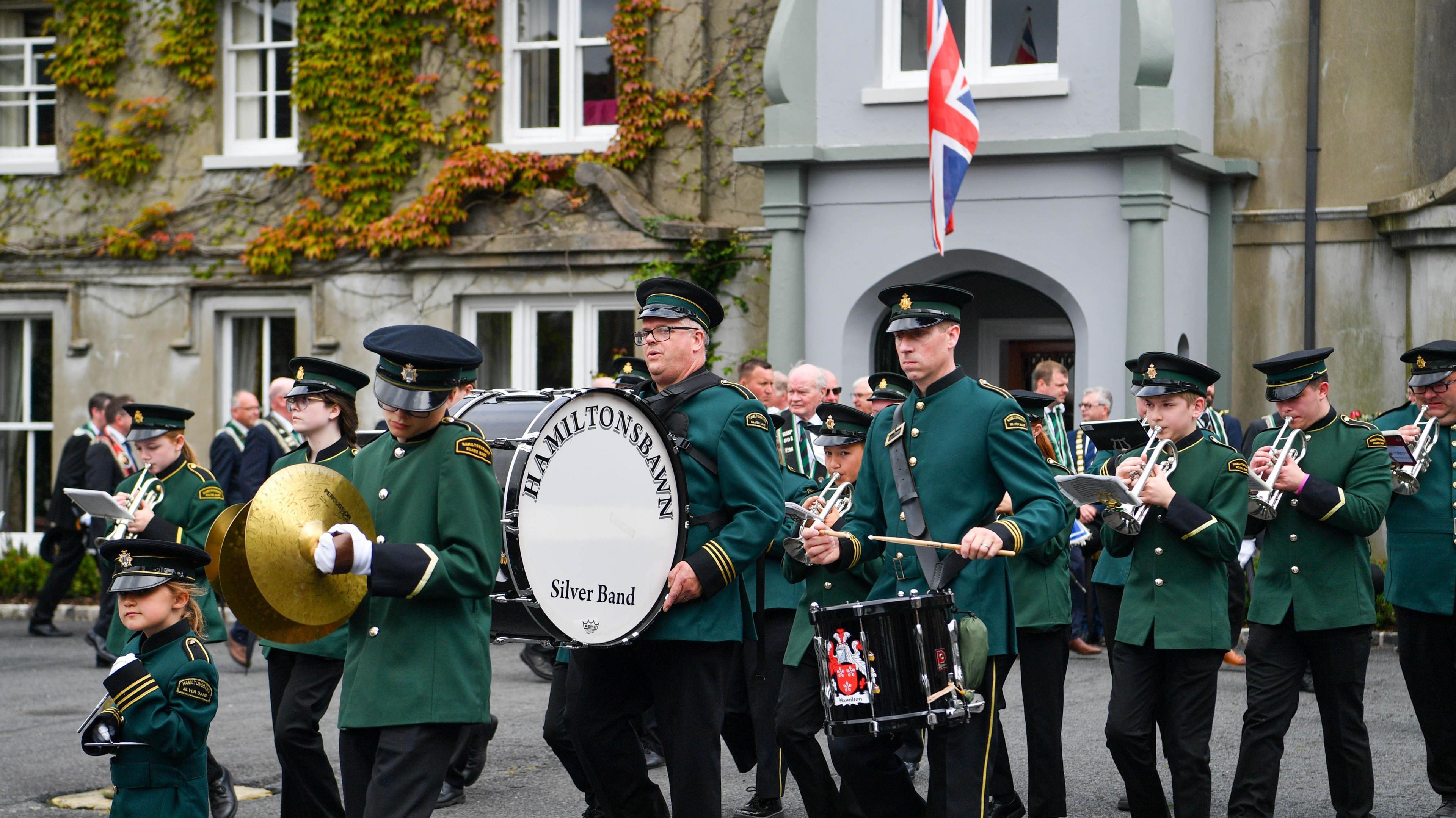 Band members wearing green uniforms and carrying drums and cymbals play outside a grey house in Scarva