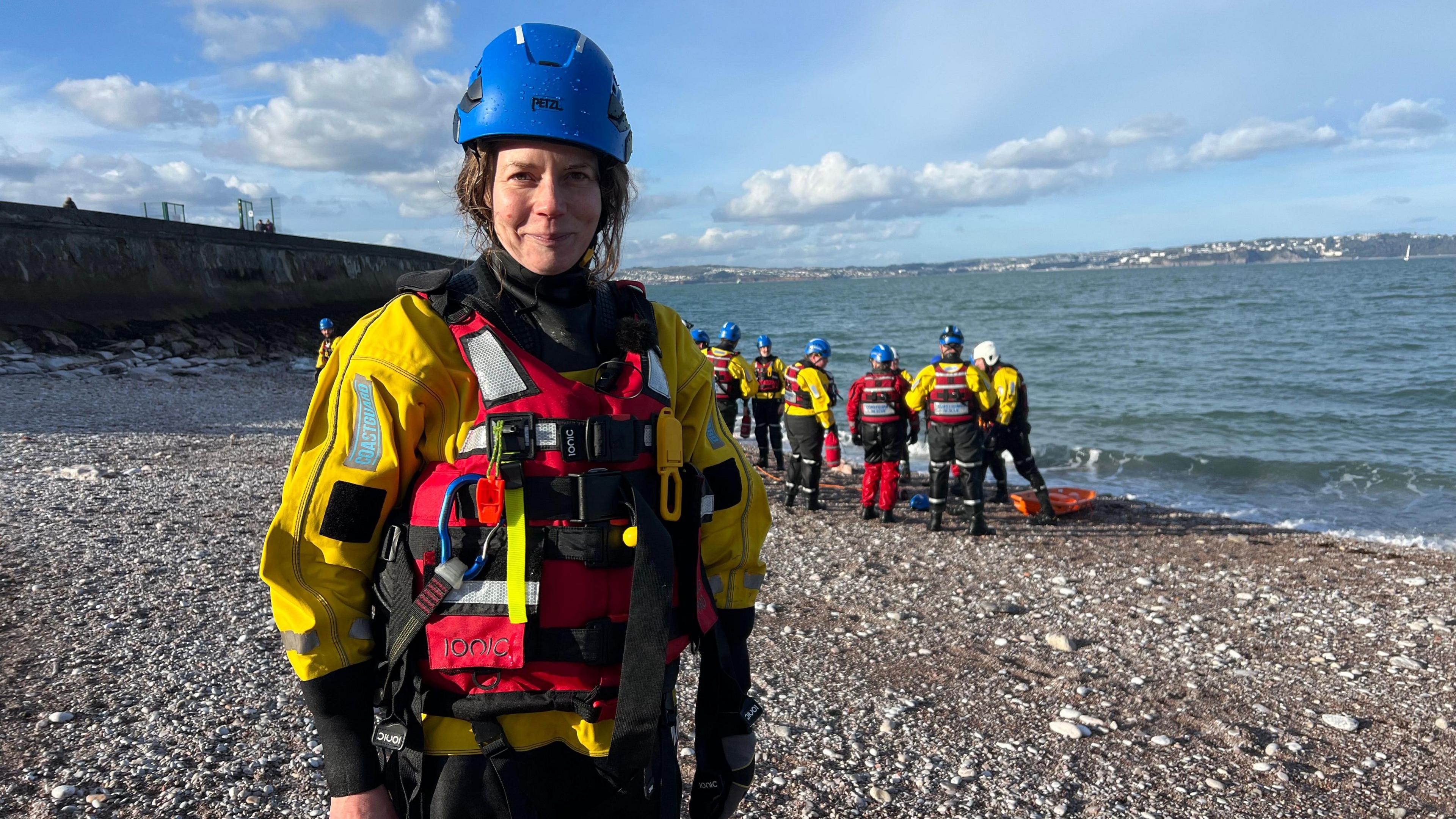 Caroline Pellow stood in her dry suit and gear at Brixham beach. She is wearing a blue hat, a red vest and yellow jacket. In the background is the other recruits near the water