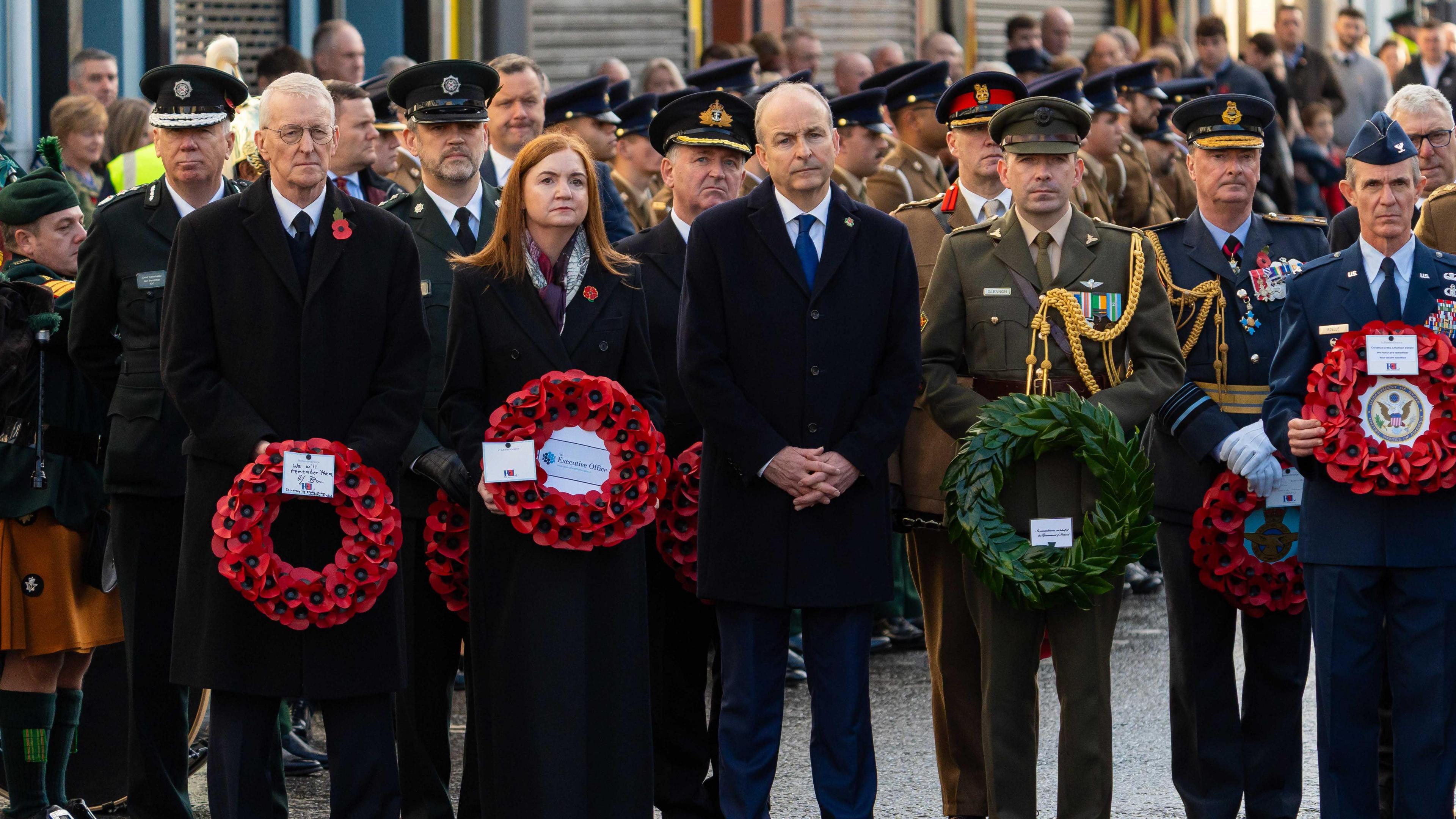 A group of politicians and people wearing army uniforms are standing close to each other. A crowd is behind them Some are holding wreths. 