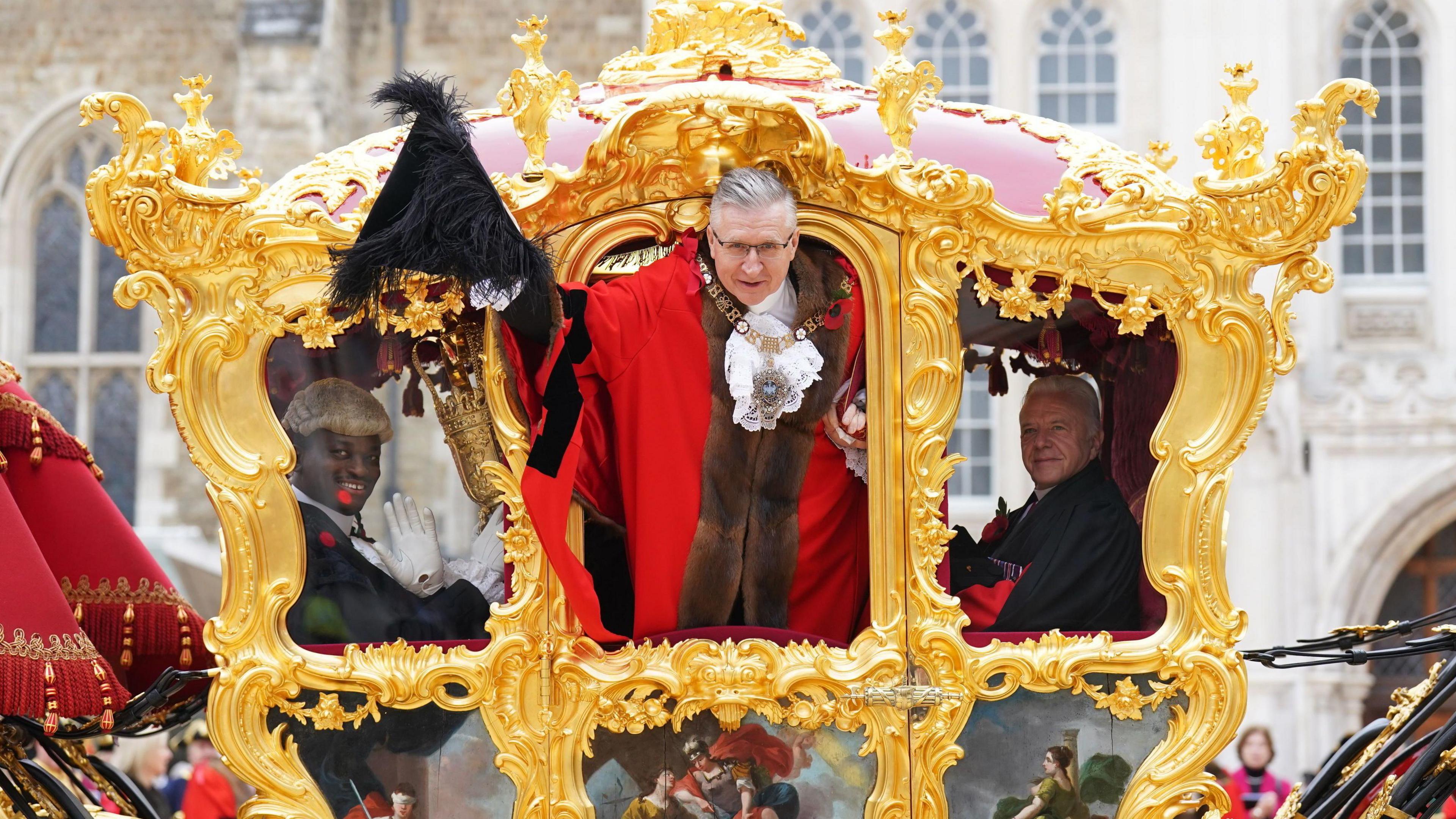 The Lord Mayor, dressed in ceremonial robes with a chain of office, leans out of a grand, gold gilded state carriage adorned with intricate decorations and paintings. He holds a black feathered hat and smiles at the crowd. Another official, also in traditional attire, sits beside him, while a another in a wig and white gloves waves from inside the carriage.