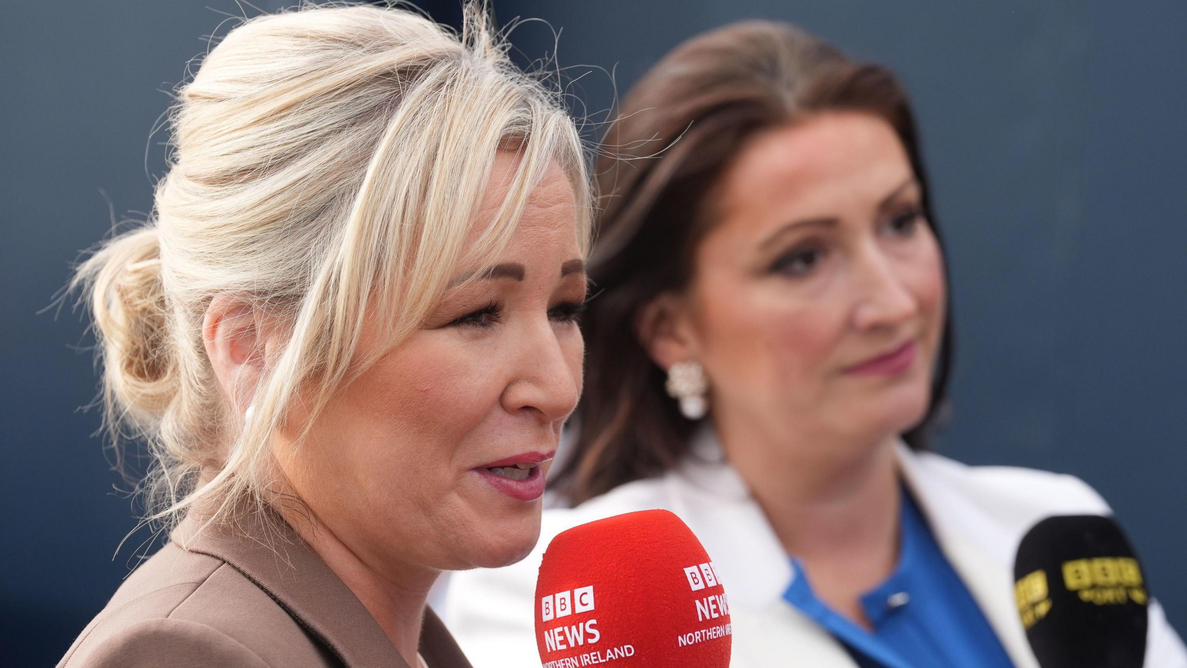Michelle O'Neill wearing a brown blazer, she is holding a red BBC News Northern Ireland microphone, she has blonde hair.
Beside her is Emma Little-Pengelly, she has black hair and is wearing a white blazer with a blue top. She is holding a black BBC Sport NI microphone.