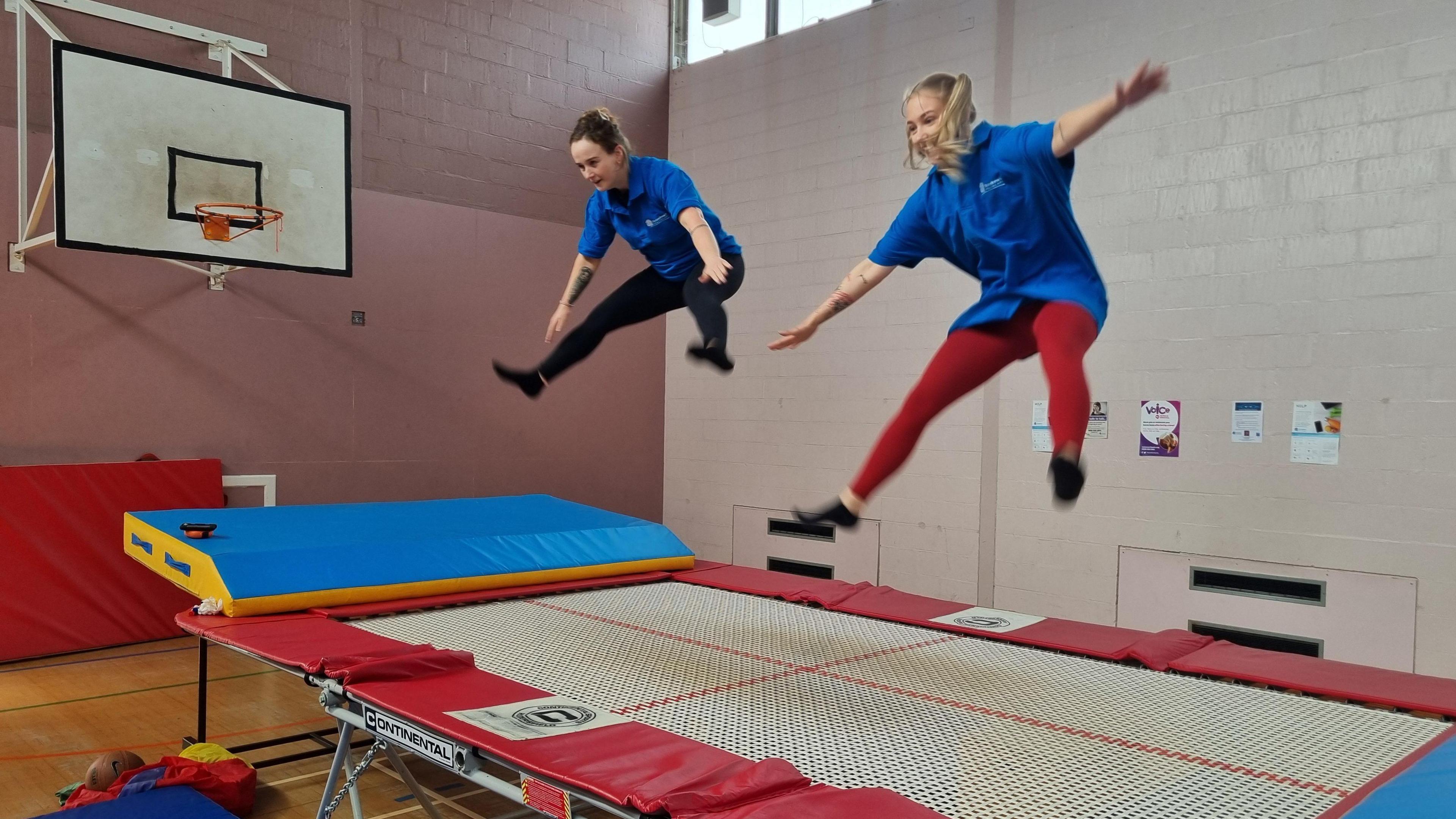 Two women in blue t-shirts mid air following a jump on a wide trampoline with red and blue trim. 