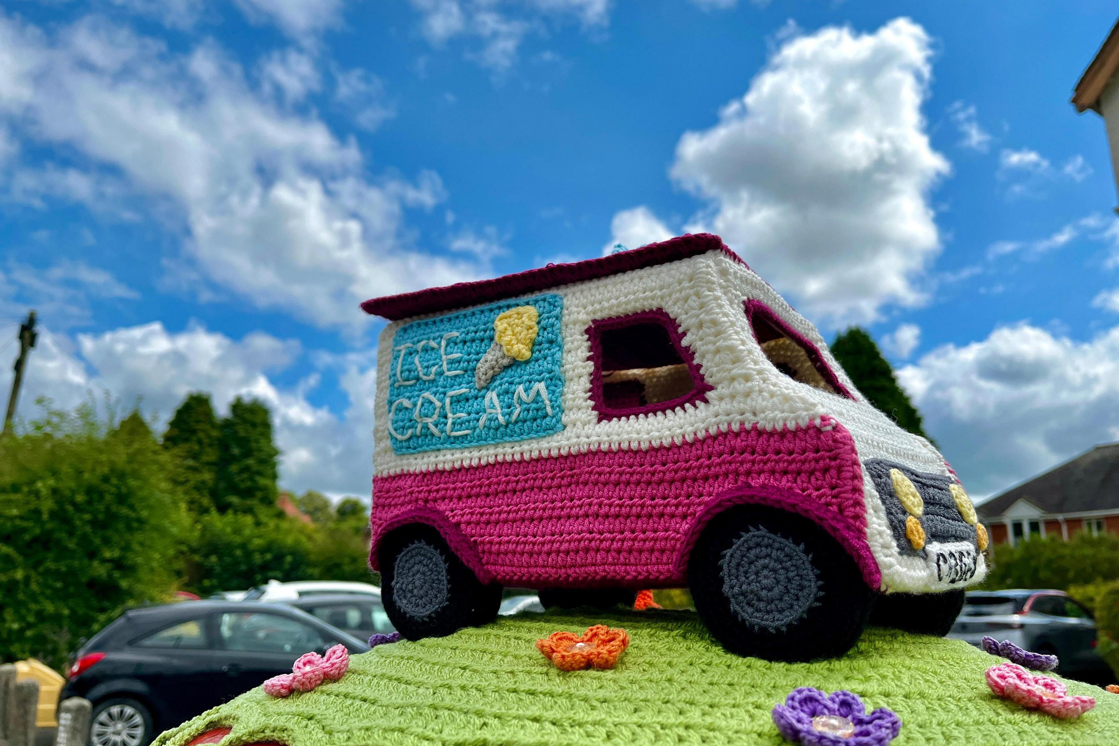 Knitted post box topper including a pick and white ice cream van atop a green field with multi-coloured flowers