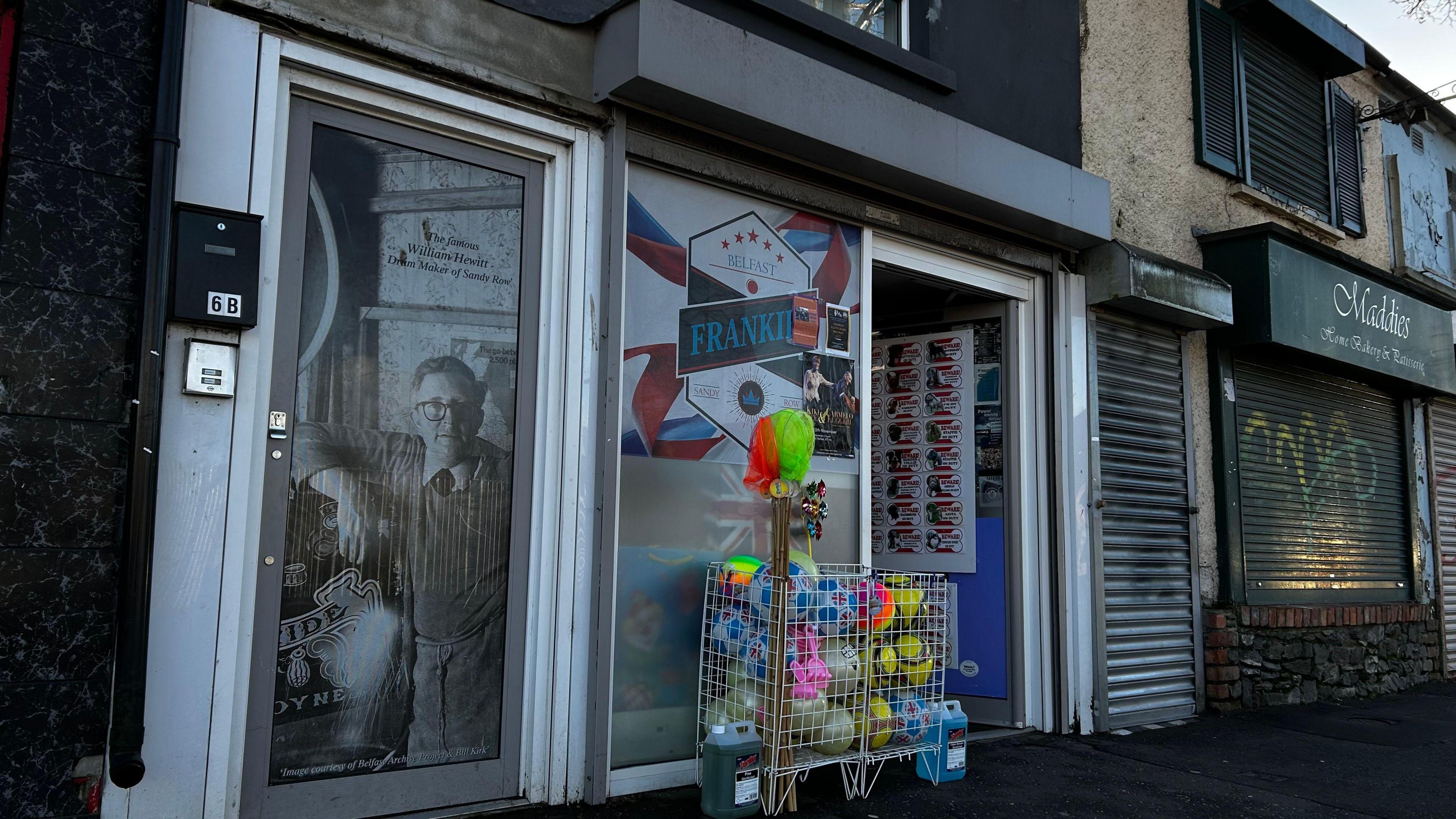 A street view of a shop front. The shop door is open and a box with football is out front. A picture of a man on a glass door can be seen to the left of the shop. A shop to the right has its shutters down.