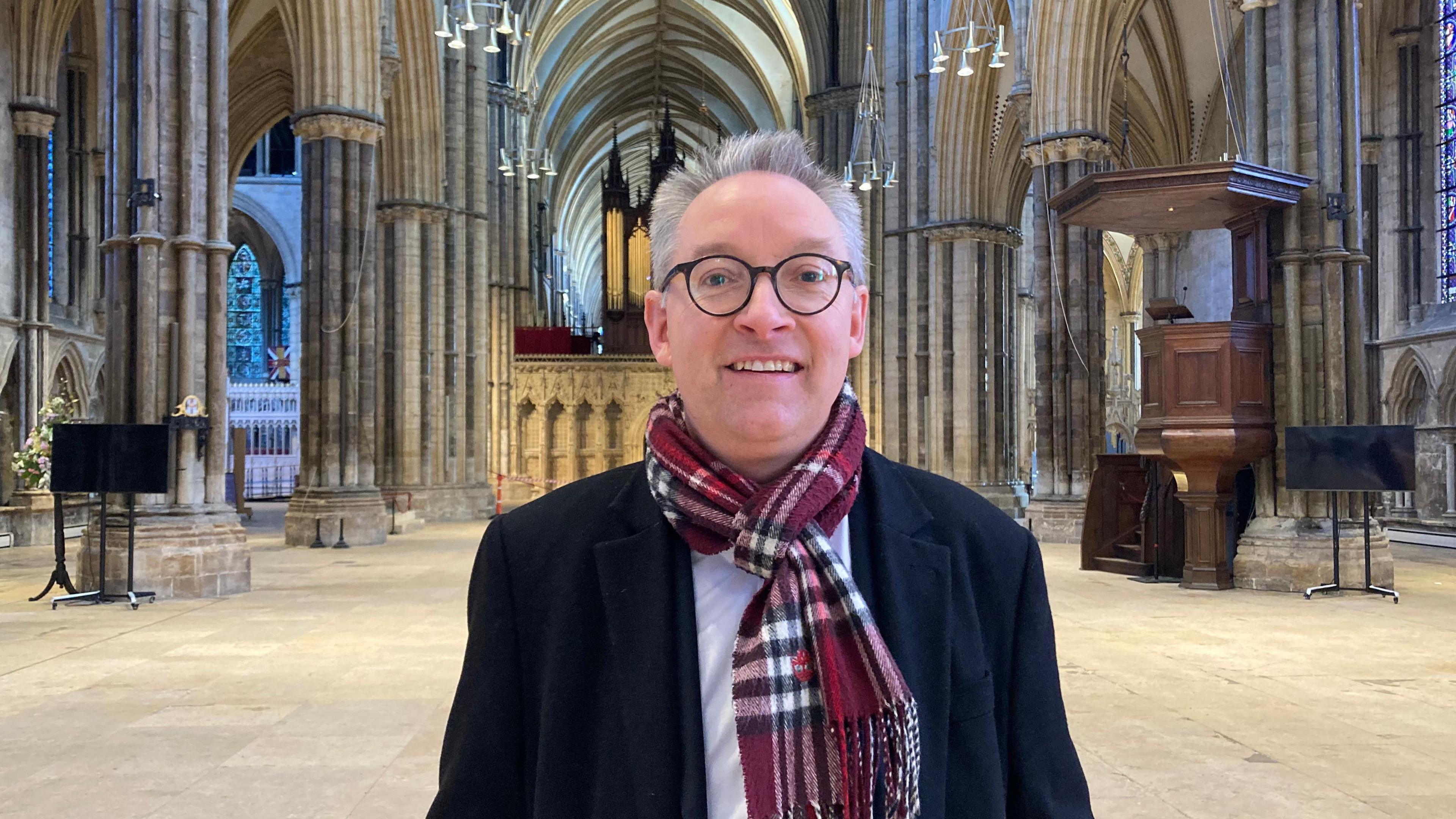 A grey-haired man, wearing glasses, a tartan scarf and a blue coat, stands in the nave of Lincoln Cathedral.