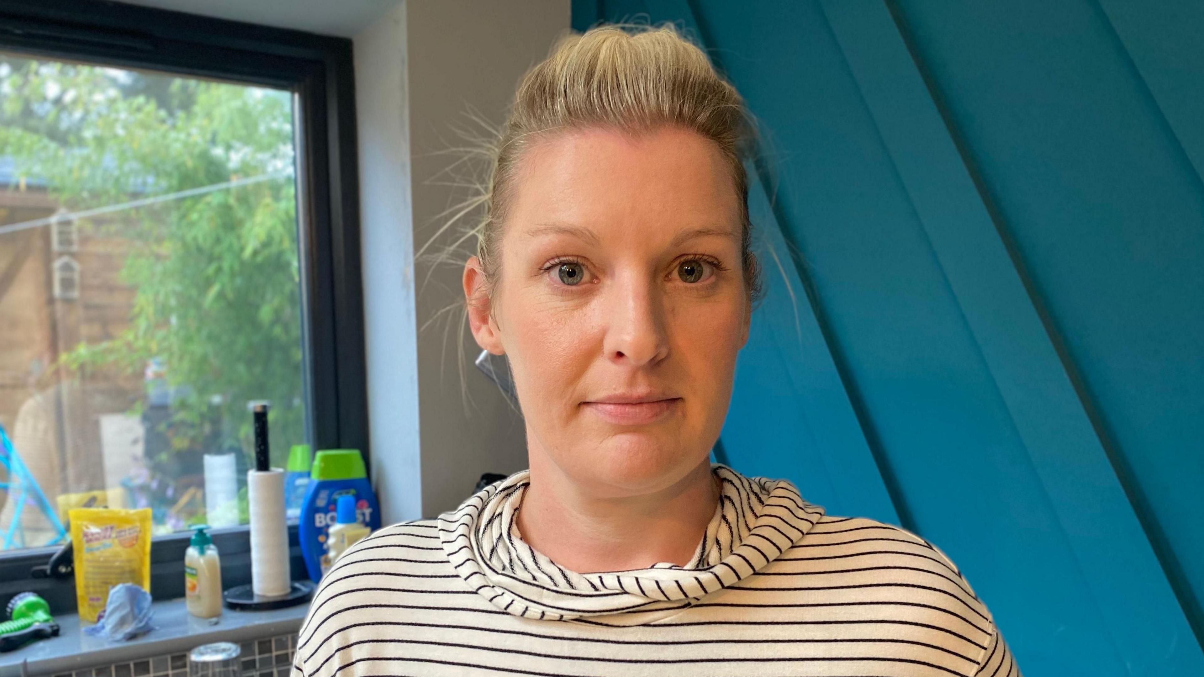 Laura Melling standing in her home, with a window to her left and a blue wall behind her. Her hair is tied back and she is a wearing a black and cream stripy top