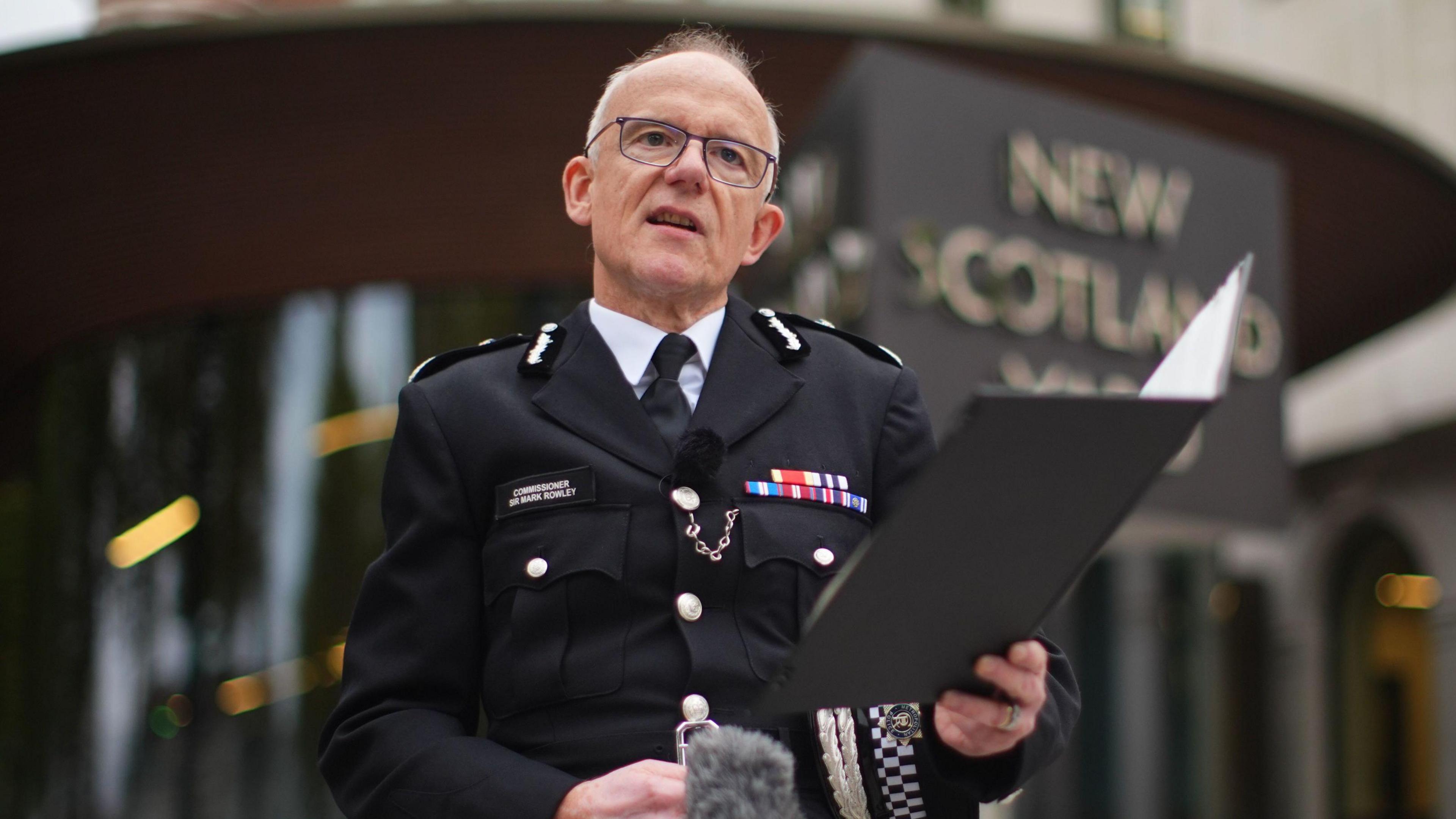 A balding middle aged man wearing police uniform stands outside New Scotland Yard police headquarters. He is reading a statement from a clipboard which he is holding in his left hand. 