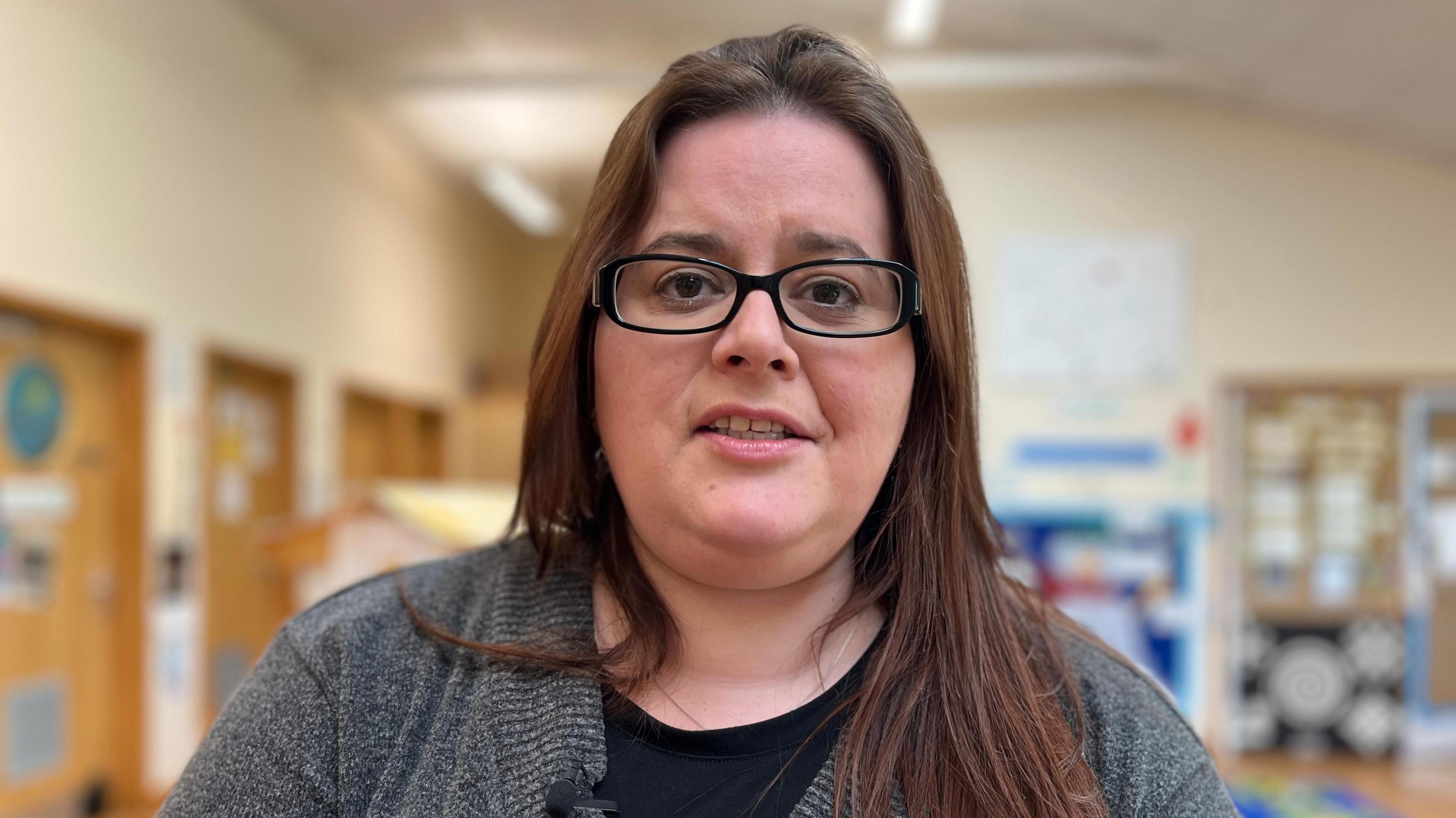 Katy Cole looks at the camera as she is photographed inside the family hub. She has long dark hair and wears black-rimmed glasses. She has a dark grey jumper and black T-shirt on. 