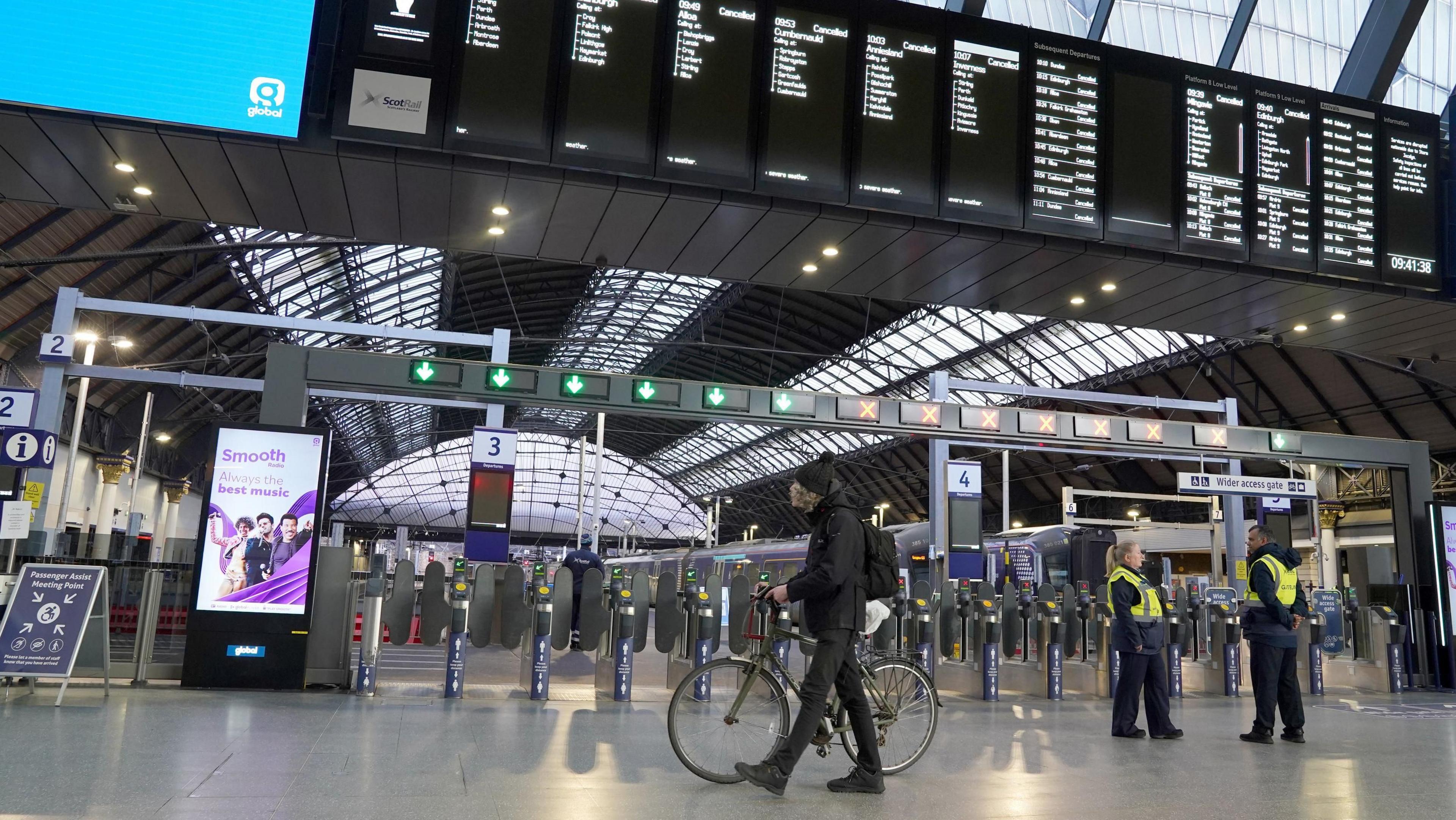 The inside of Glasgow's Queen Street railway station in January 2024, when trains were cancelled due to Storm Jocelyn. A man dressed in black is pushing a bike and two staff wearing high vis jackets are standing nearby. The station is otherwise empty and the departure board shows that services are canncelled.