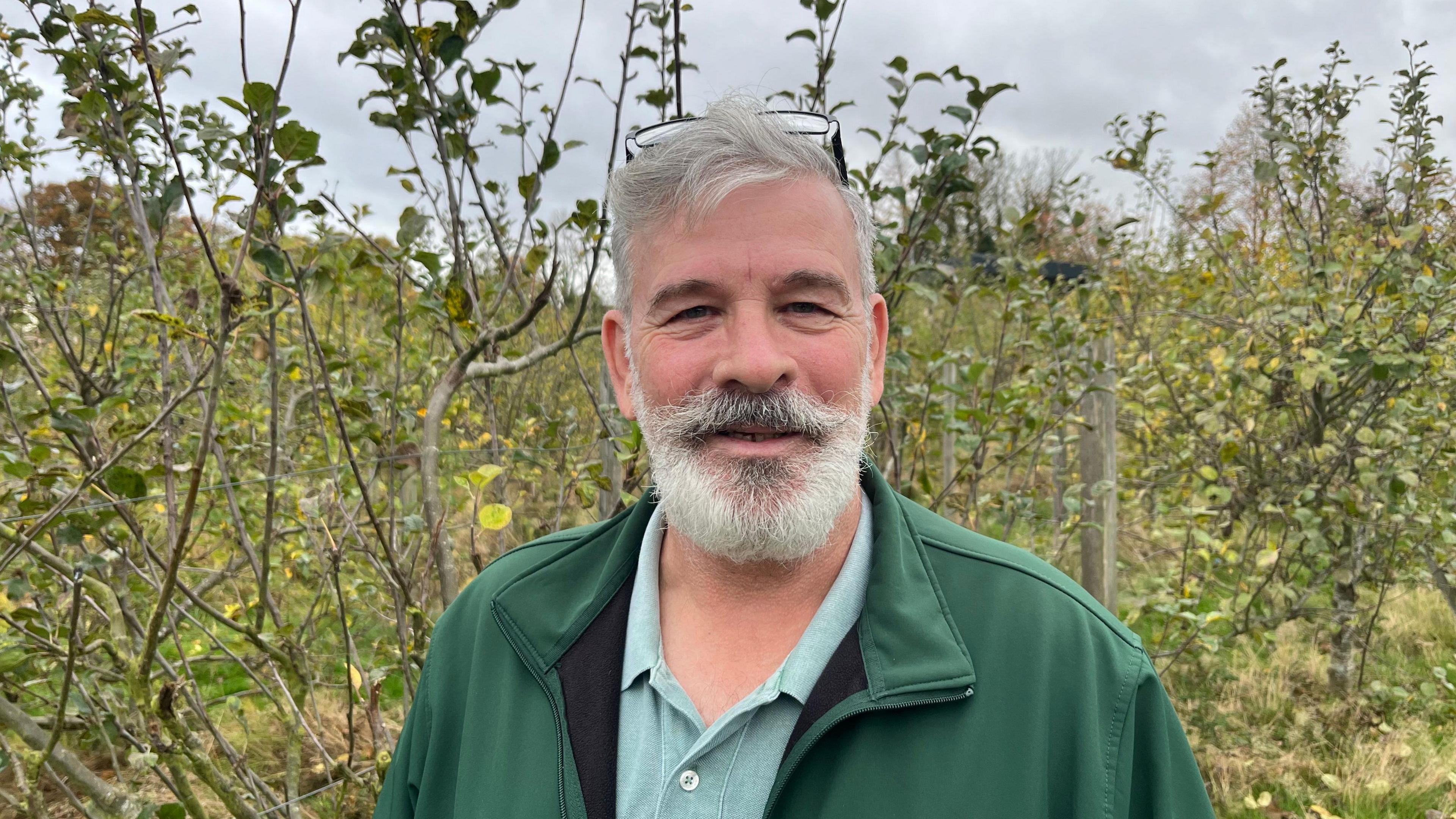 Andrew Callaghan, 60, has a grey beard and moustache and matching hair, he is wearing a green fleece top over light green t-shirt with a buttoned collar.
Behind him is part of the apple orchard.
