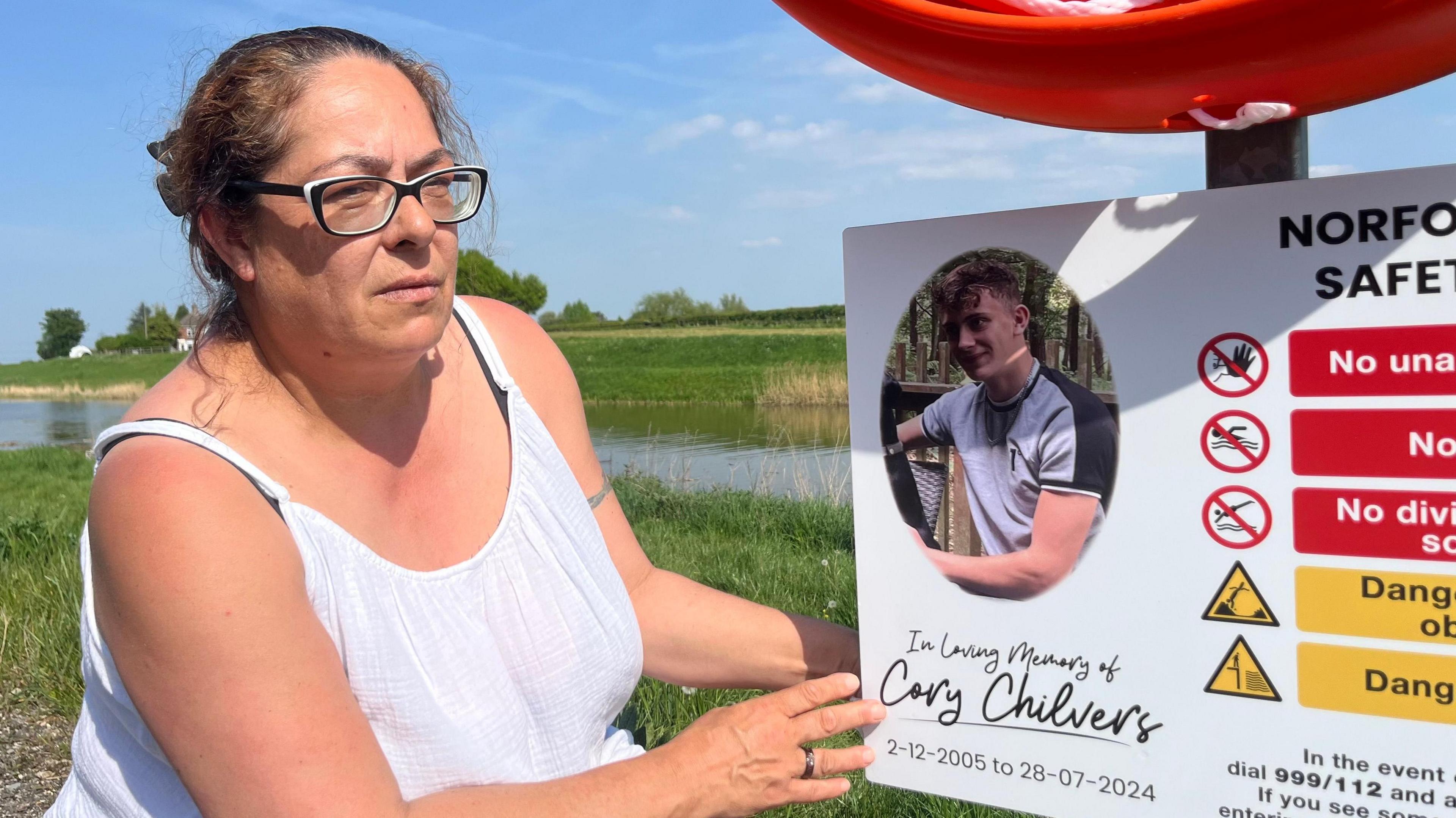 Claire Wadham stands next to the safety sign with a picture of her son Cory Chilvers who drowned in water at the site of the sign in July 2024.