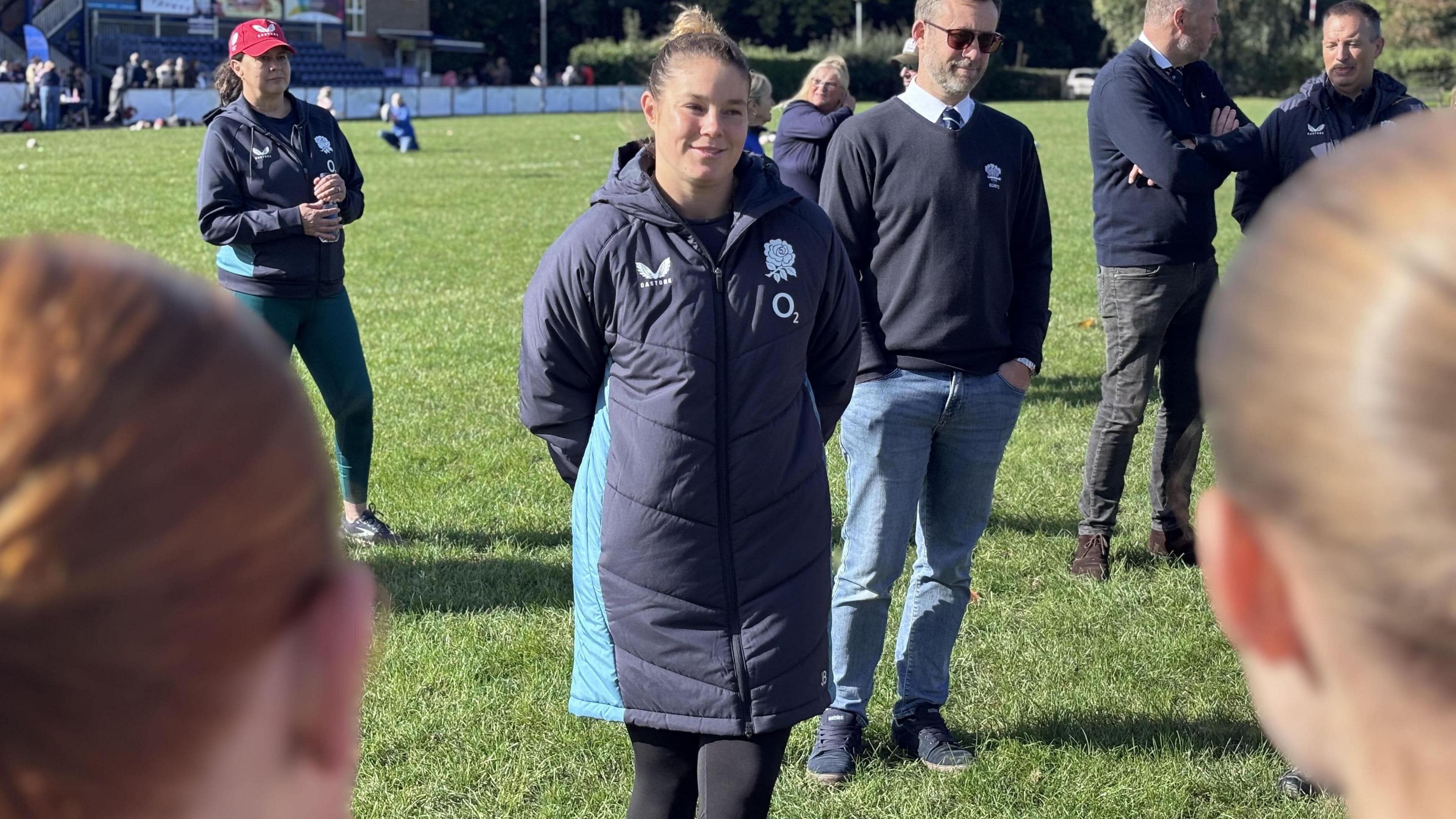 Jess Breach is standing on a rugby pitch and wearing a long training jacket bearing the famous England rose. She is smiling at fans who have turned out to greet her. 