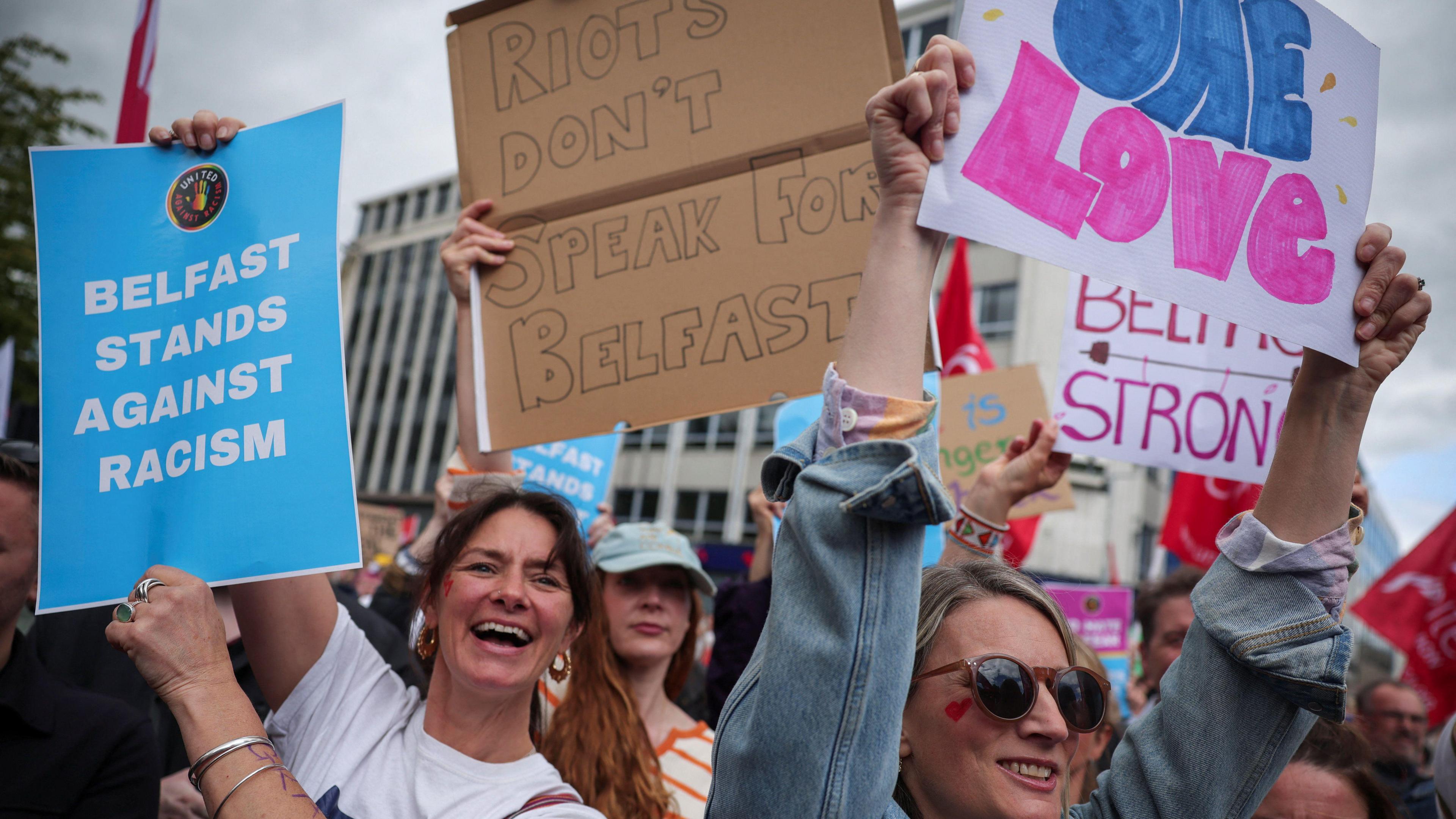 Women hold up signs saying "Belfast stands against racism" "One Love", and "Riots don't speak for Belfast". They are in an urban area, and there are grey clouds above them. 