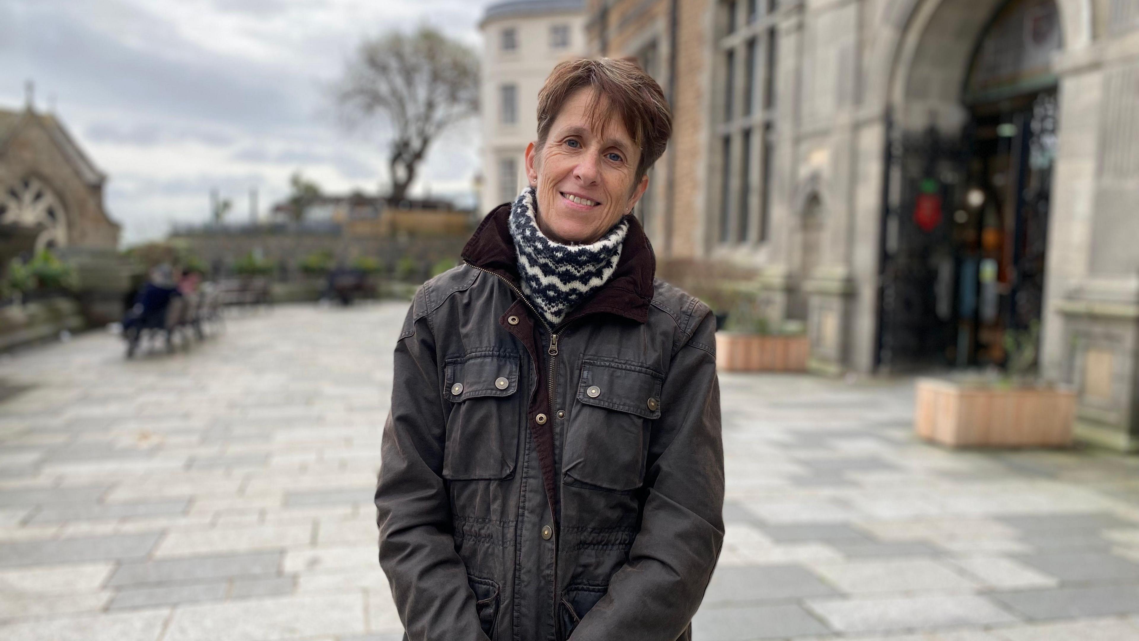 A woman with short brown hair in a brown jacket and white scarf stands in Guernsey's Market Square.