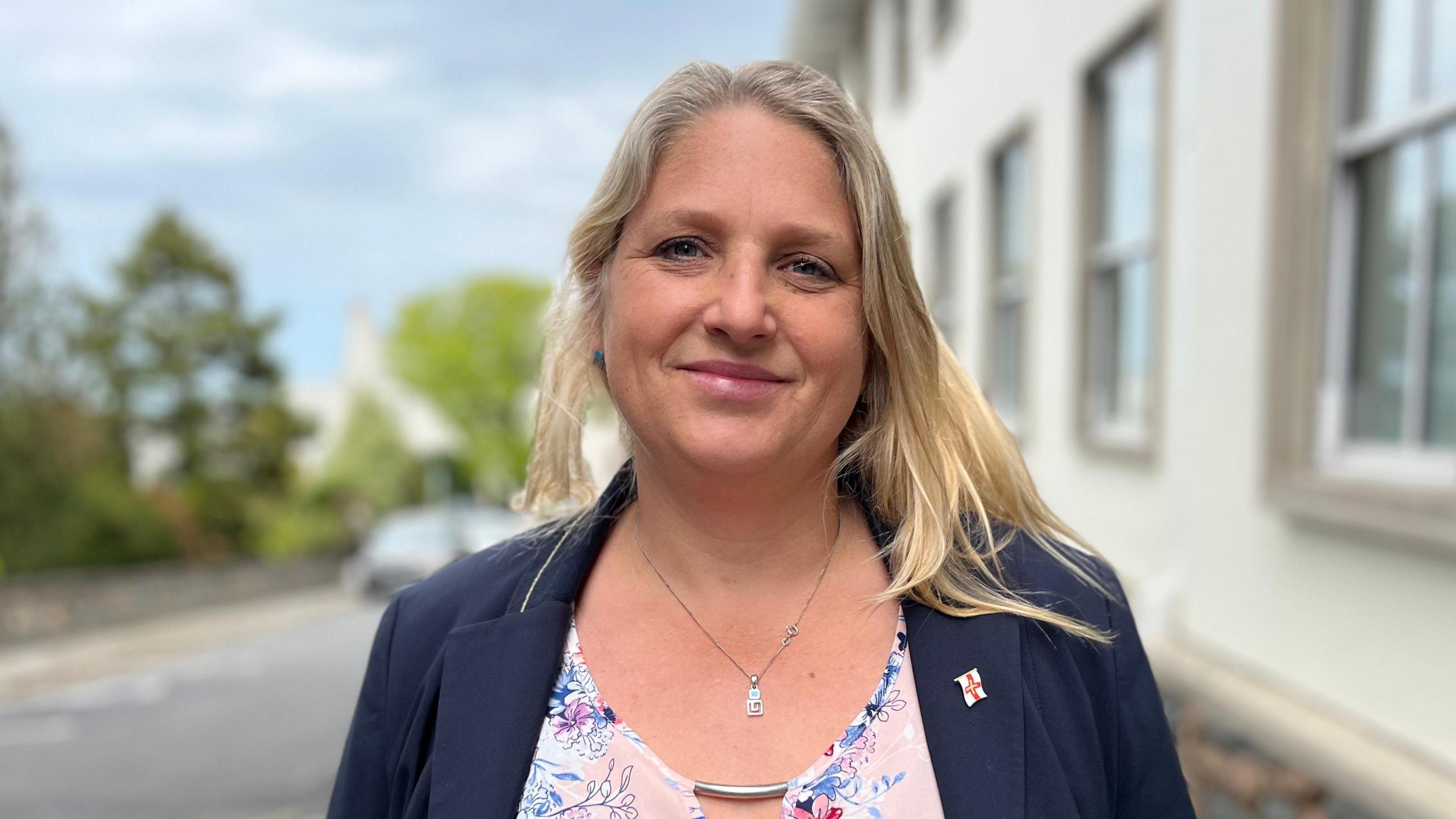 Deputy Victoria Oliver, who has blonde hair, smiling at the camera wearing a blue jumper with a Guernsey pin badge and a flowery pink blouse. 