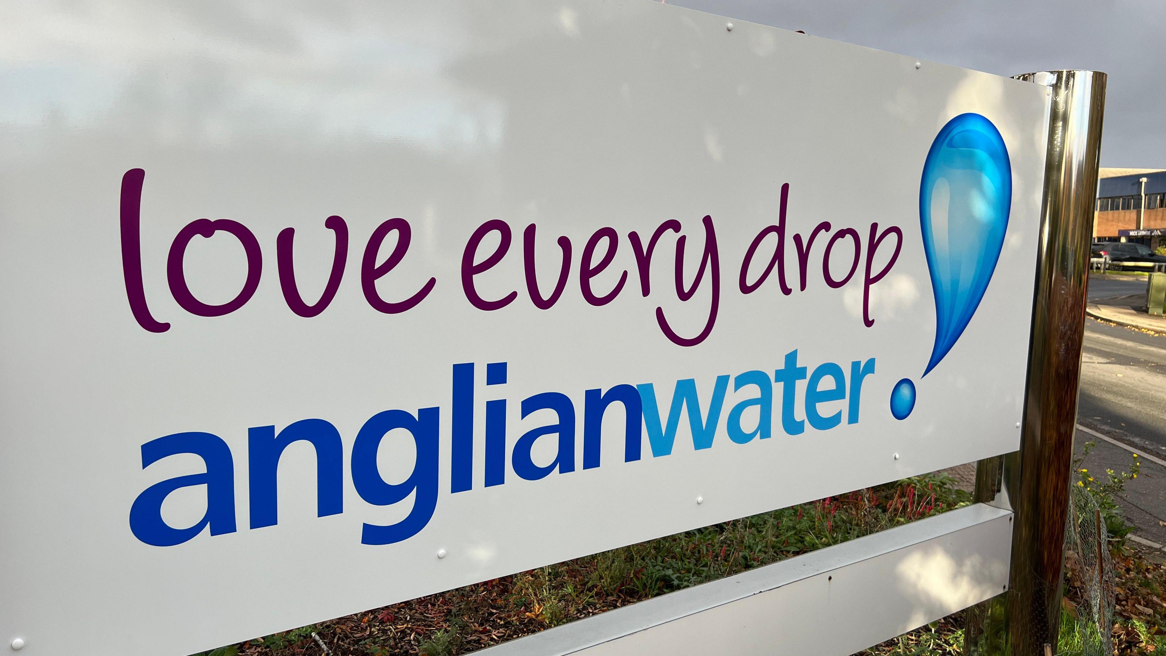 A sign spans across the whole of the image. It is white, and has in blue and purple writing, 'Love every drop Anglian Water.' In the background, there is a building and a road. 