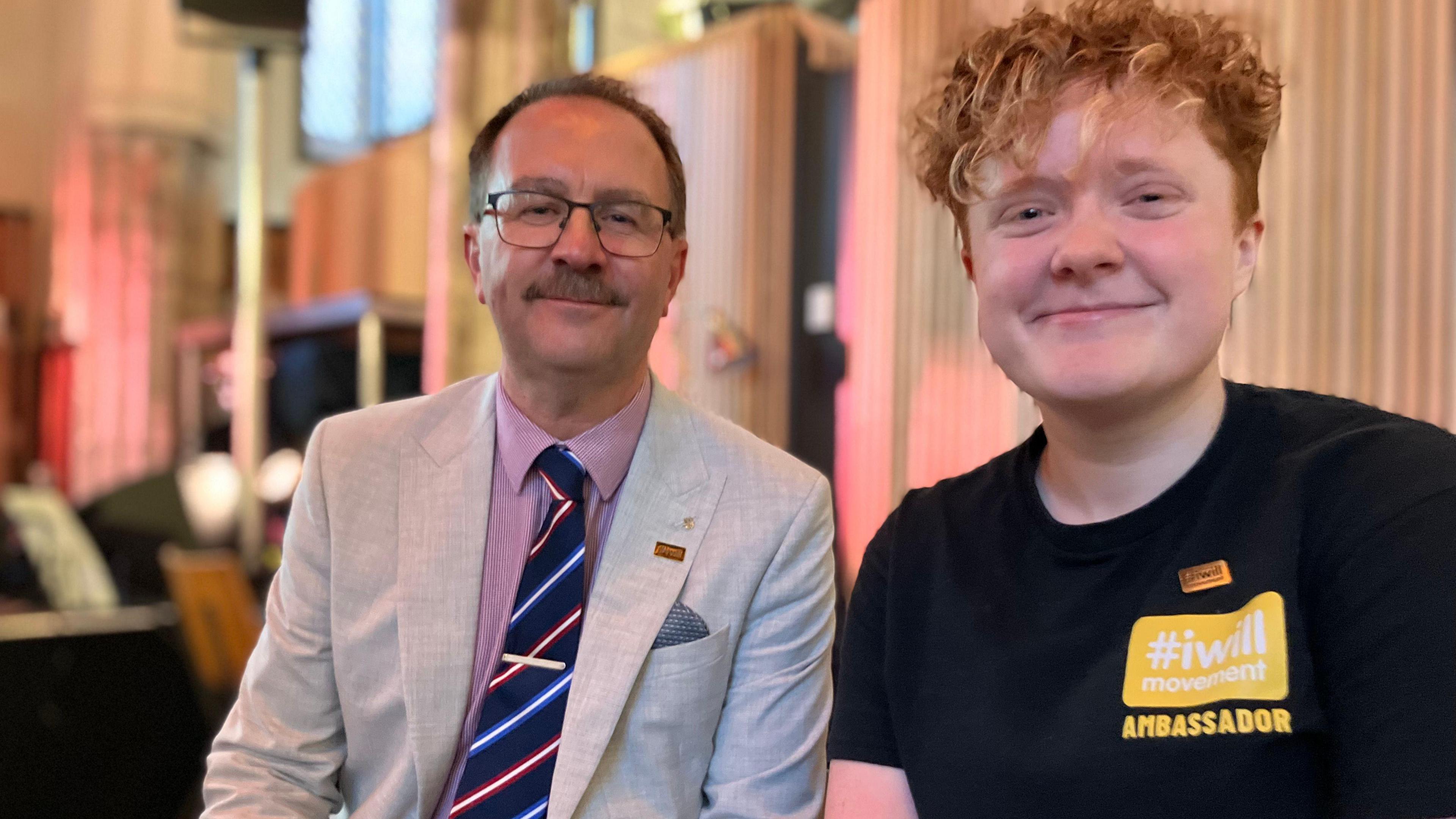 A man in a jacket and tie, and a young person in an Iwill t-shirt smile at the camera.
