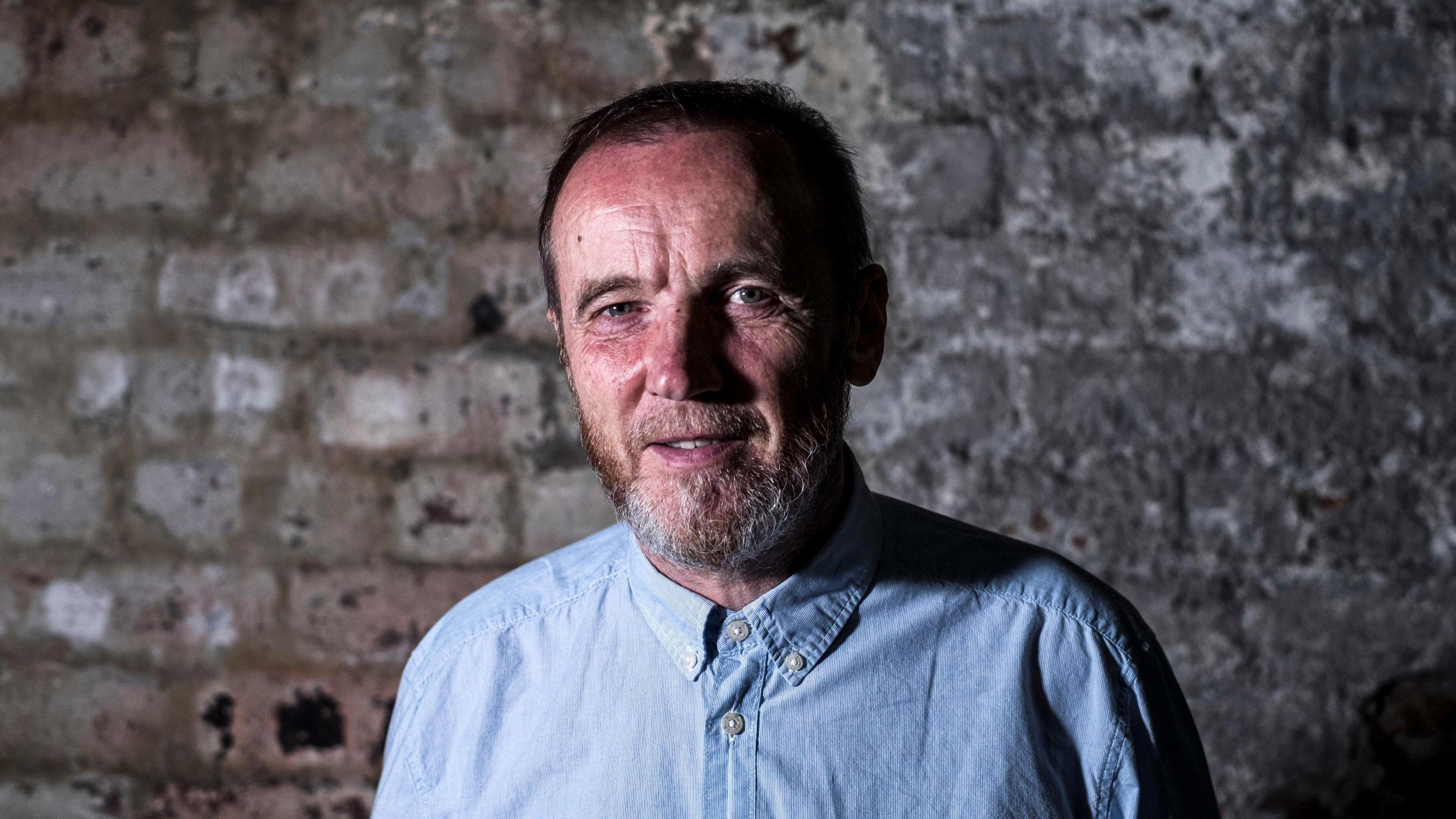 A man with a greying beard and a blue shirt smiles for the camera while standing indoors