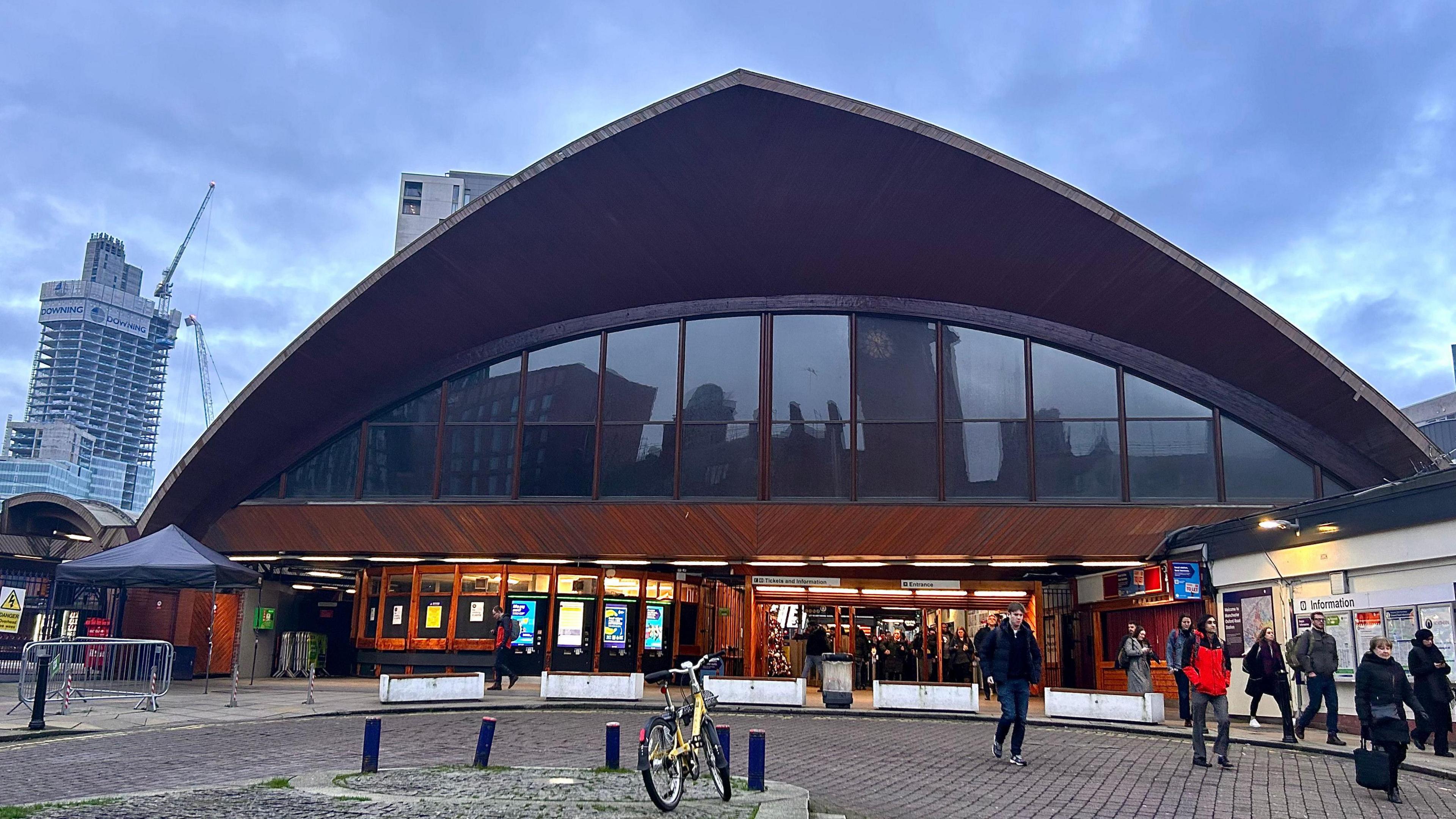 The timber facade of Manchester Oxford Road looms above the entrance to the station. Passengers can be seen walking out from the ticket stiles below on to a paved concourse. Several high-rise buildings can be seen in the distance on a cloudy day.