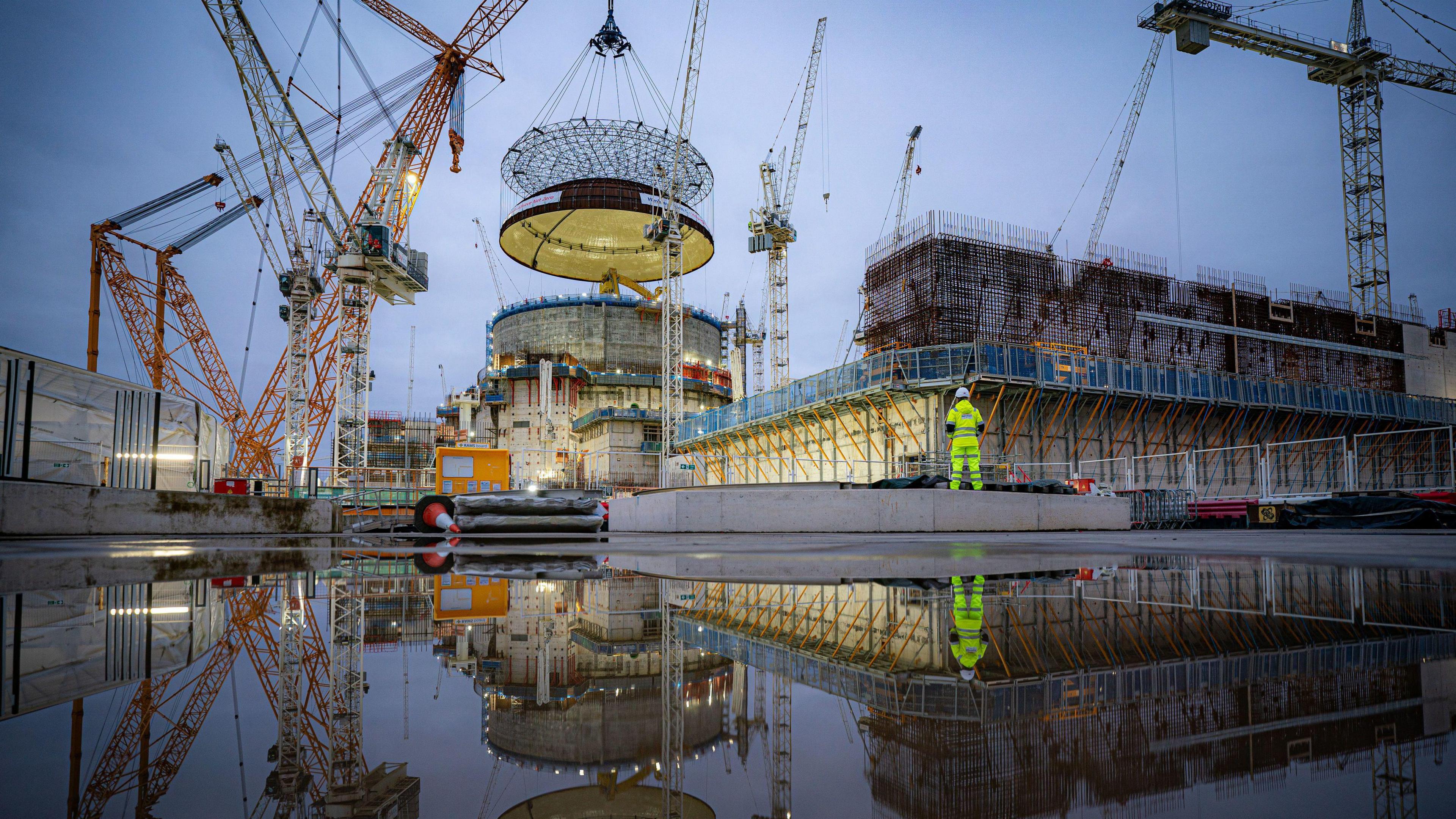 Engineering teams use the world's largest crane - Big Carl - to lift a 245-tonne steel dome onto Hinkley Point C's first reactor building, at the nuclear power station construction site in Bridgwater, Somerset. The scene is reflected in water below it, including a construction worker in high-vis standing at the front of the buildings.