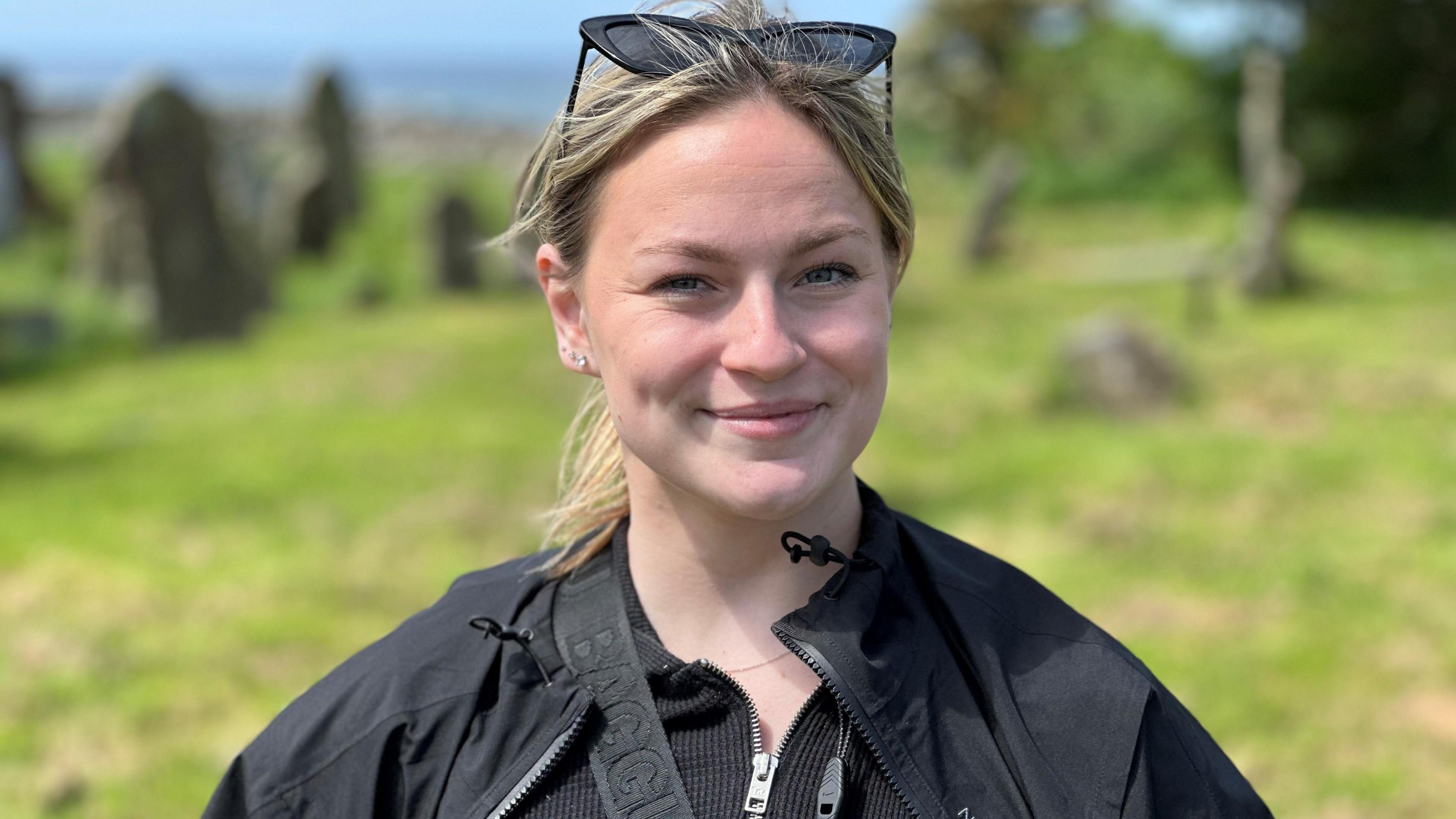 A young woman standing in a churchyard. She has long blonde hair tied back in a ponytail, is wearing a black jacket and has sunglasses on her head.