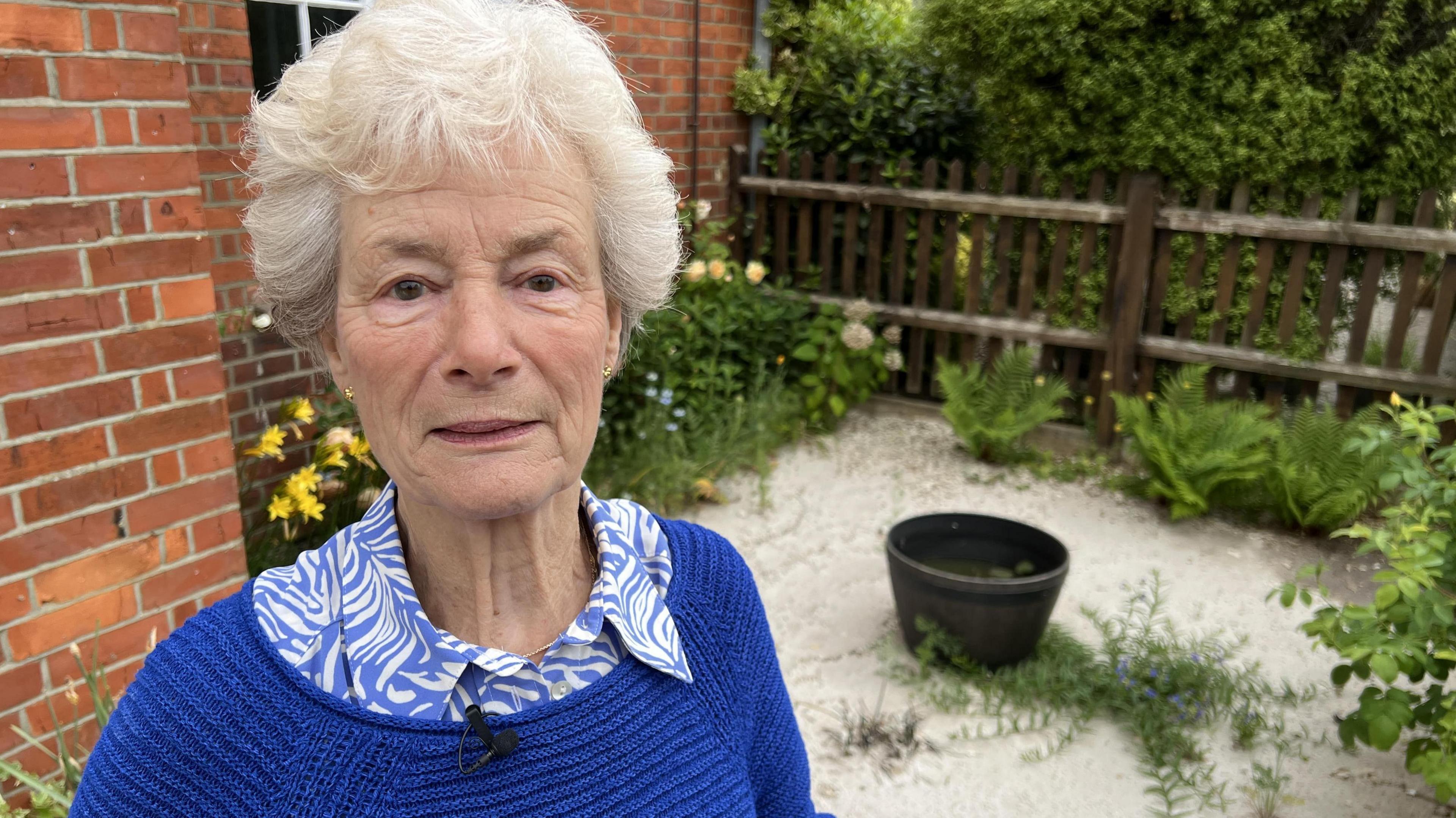 A woman with white hair looks into camera with her damaged garden behind her