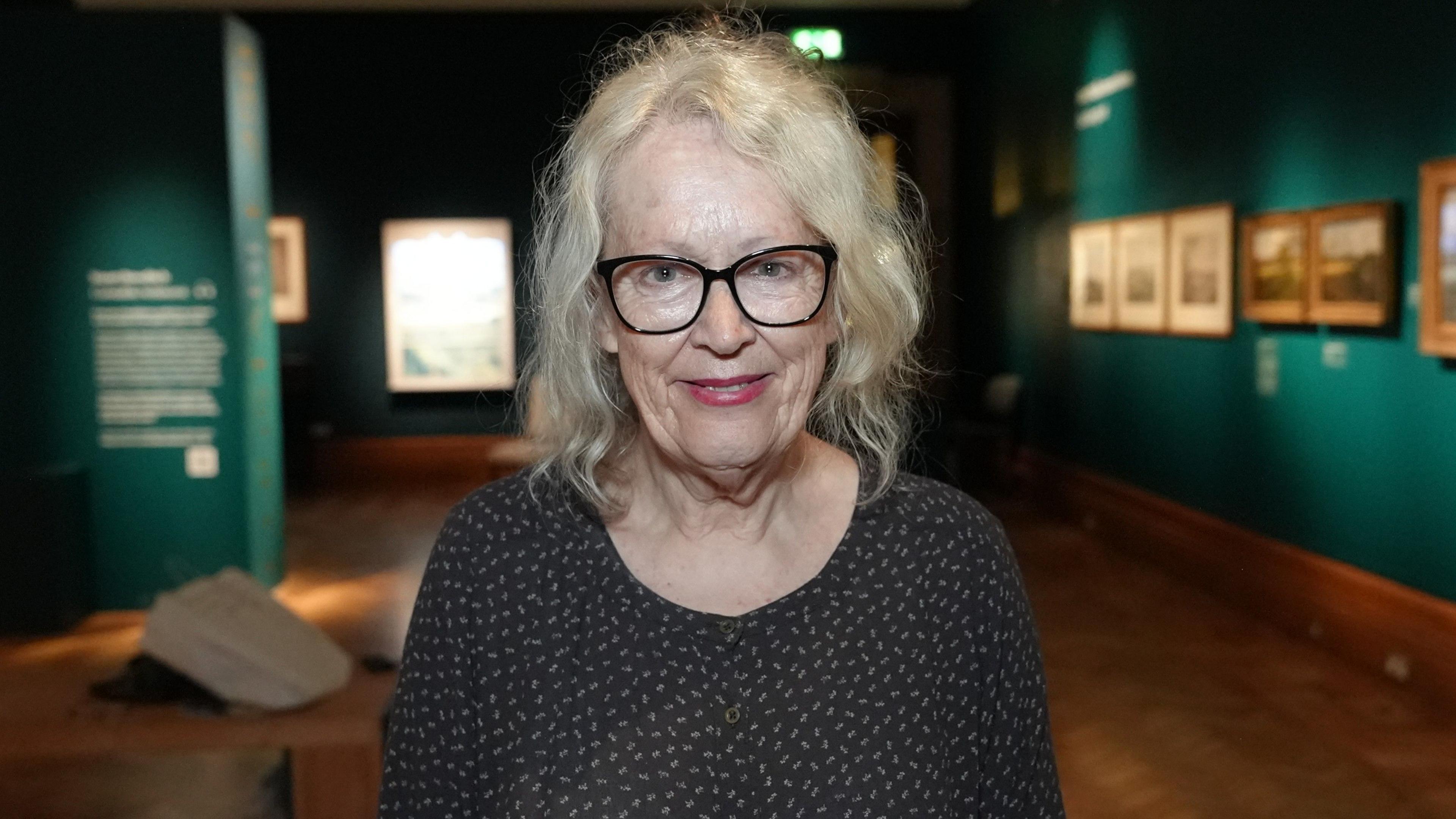 A woman standing in the middle of an art exhibition. She has shoulder length grey hair and has glasses as well as a black top with some white specks on it. 
