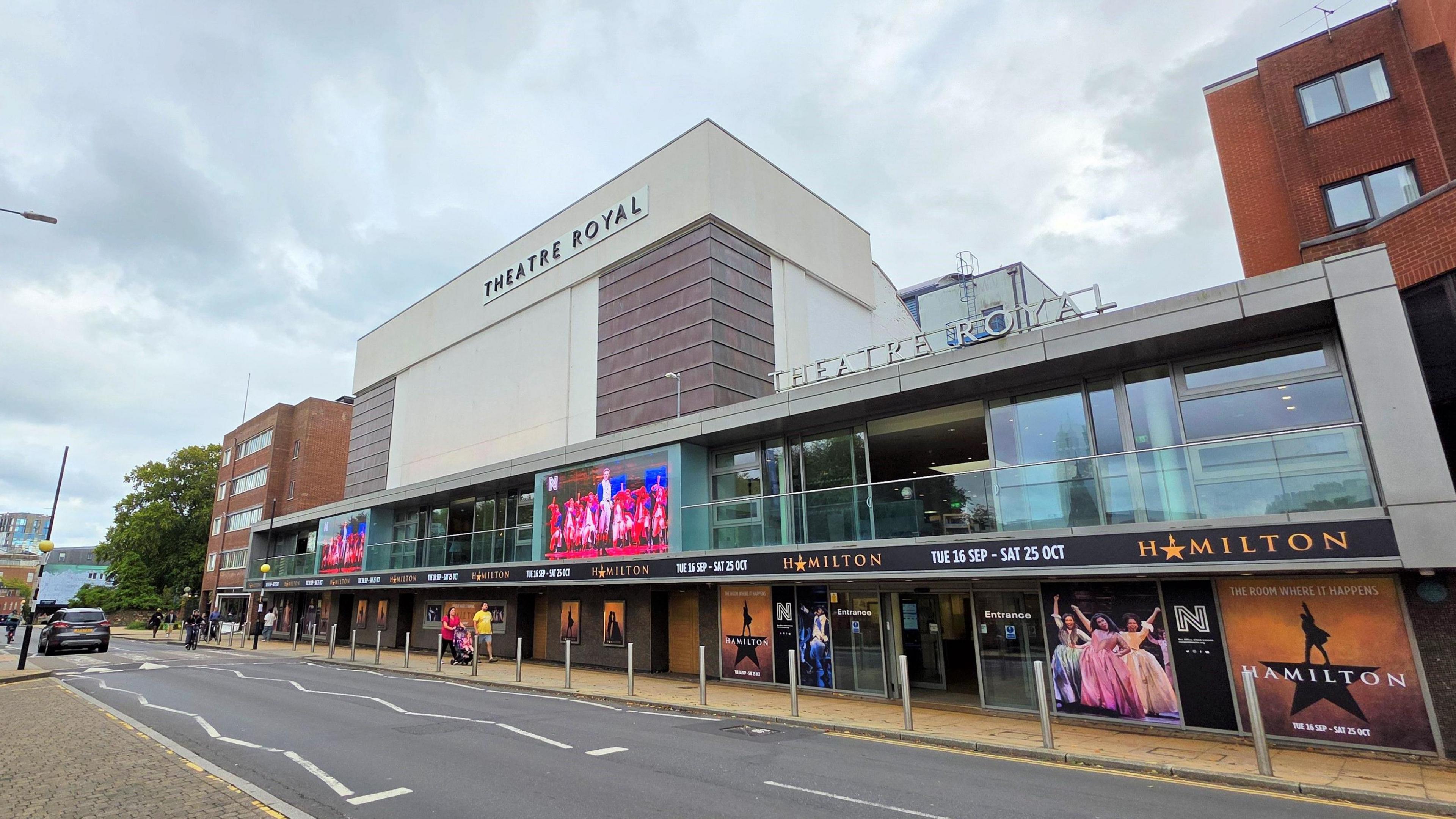 We can see the front of Norwich Theatre Royal, advertising the show 'Hamilton'. The photo is taken from across the road. There are clouds above.