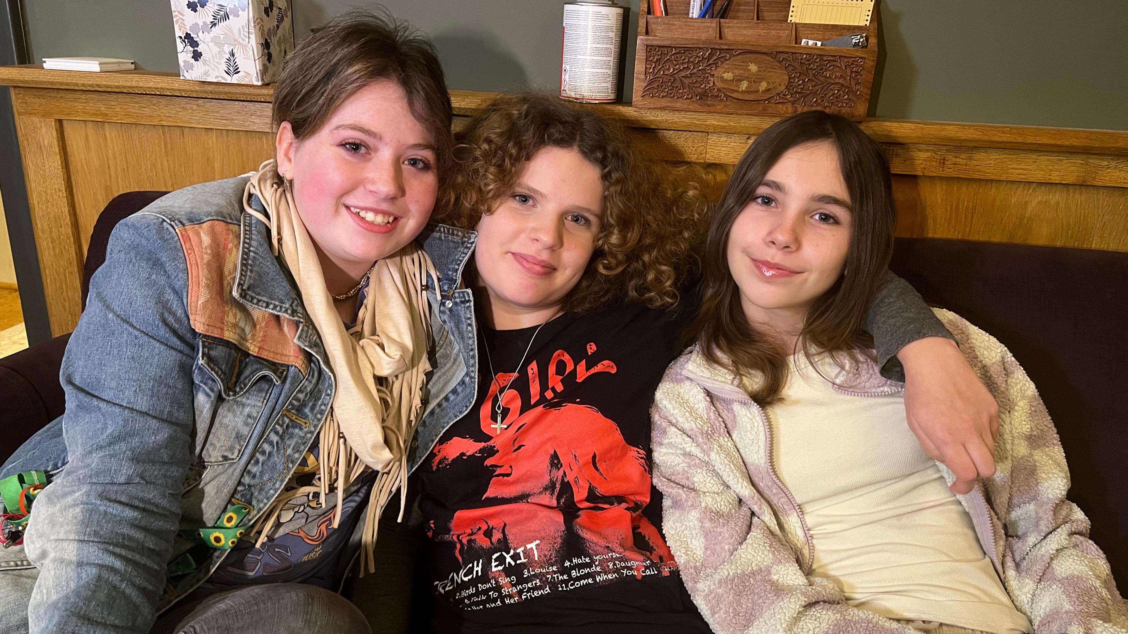 Jess, Alice and Ines (left to right) sit together on a sofa in the Cheese and Grain studio. They are putting their arms around each other and smiling at the camera.