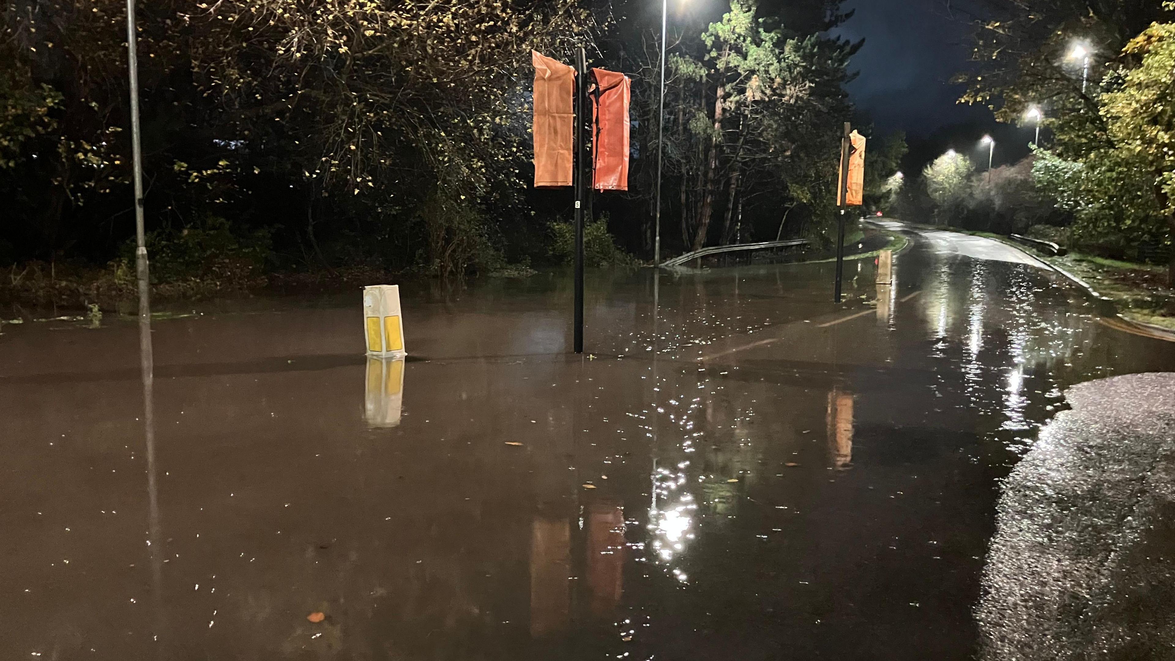 Water flooding the road on Piper's Way, which is empty of cars. It is dark and the streetlights are reflecting on the water. 