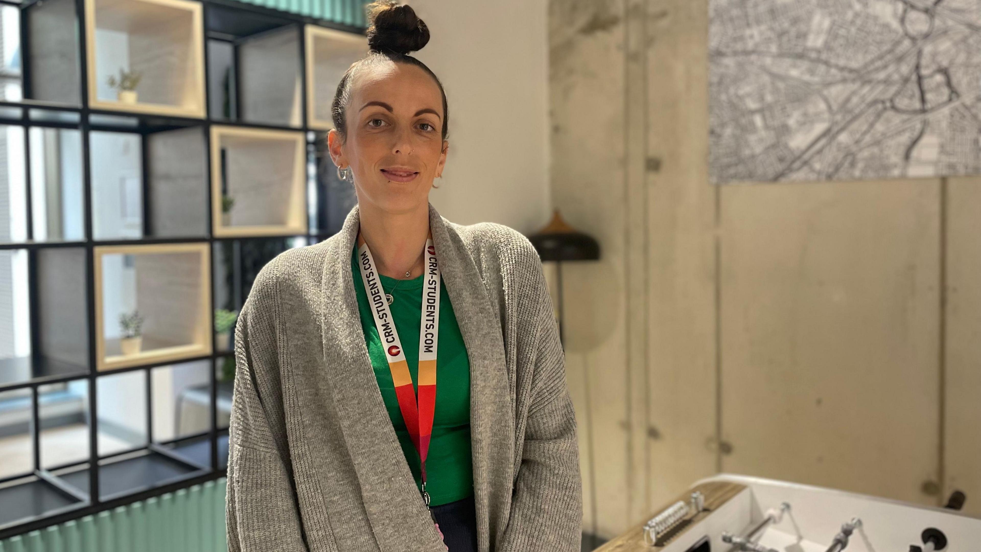 A woman wearing a grey cardigan and multicoloured lanyard looks at the camera. She has brown hair tied up on top of her head and silver hoop earrings.